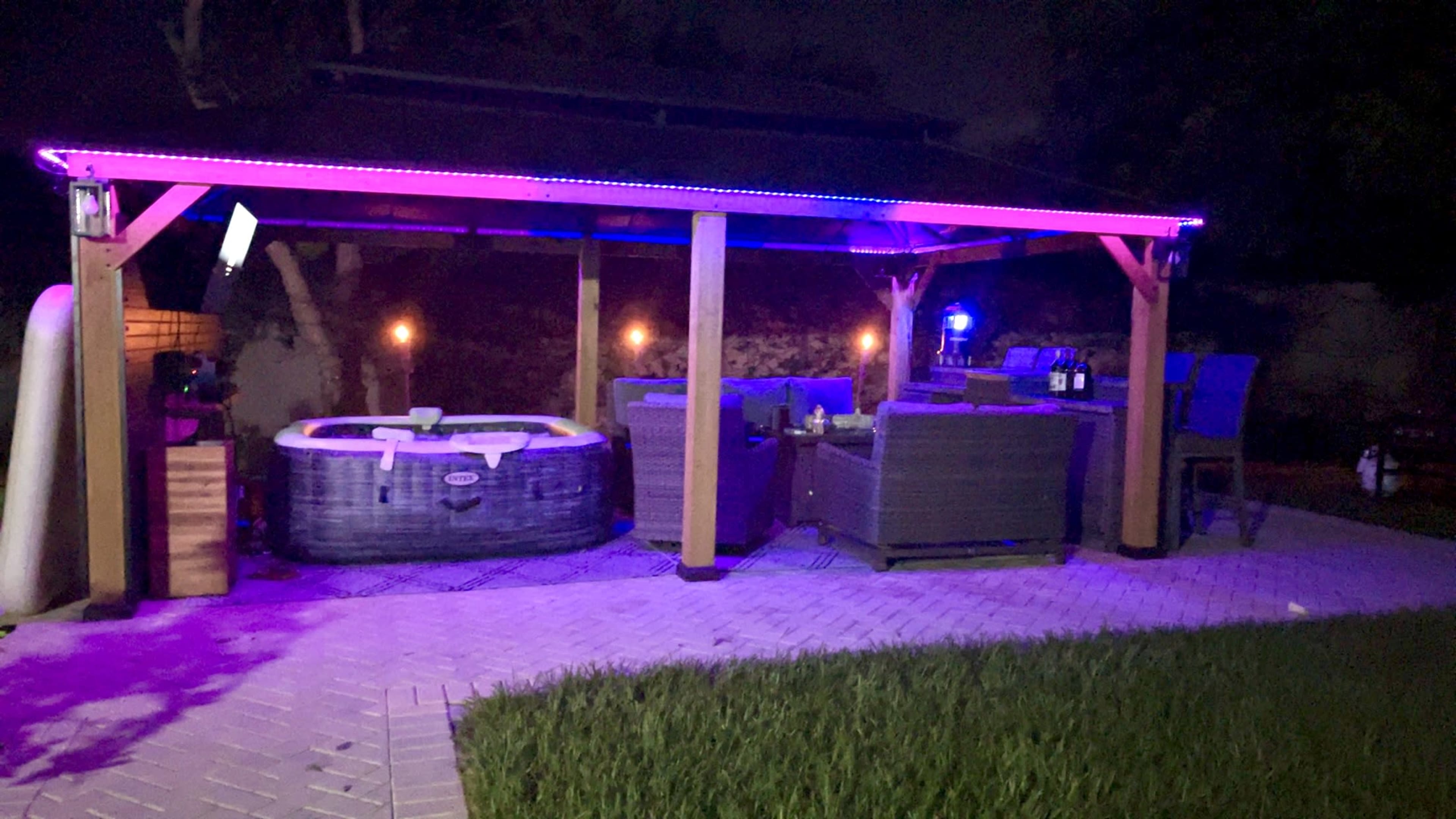 A covered outdoor patio features a hot tub and seating area illuminated by colored lights at night.