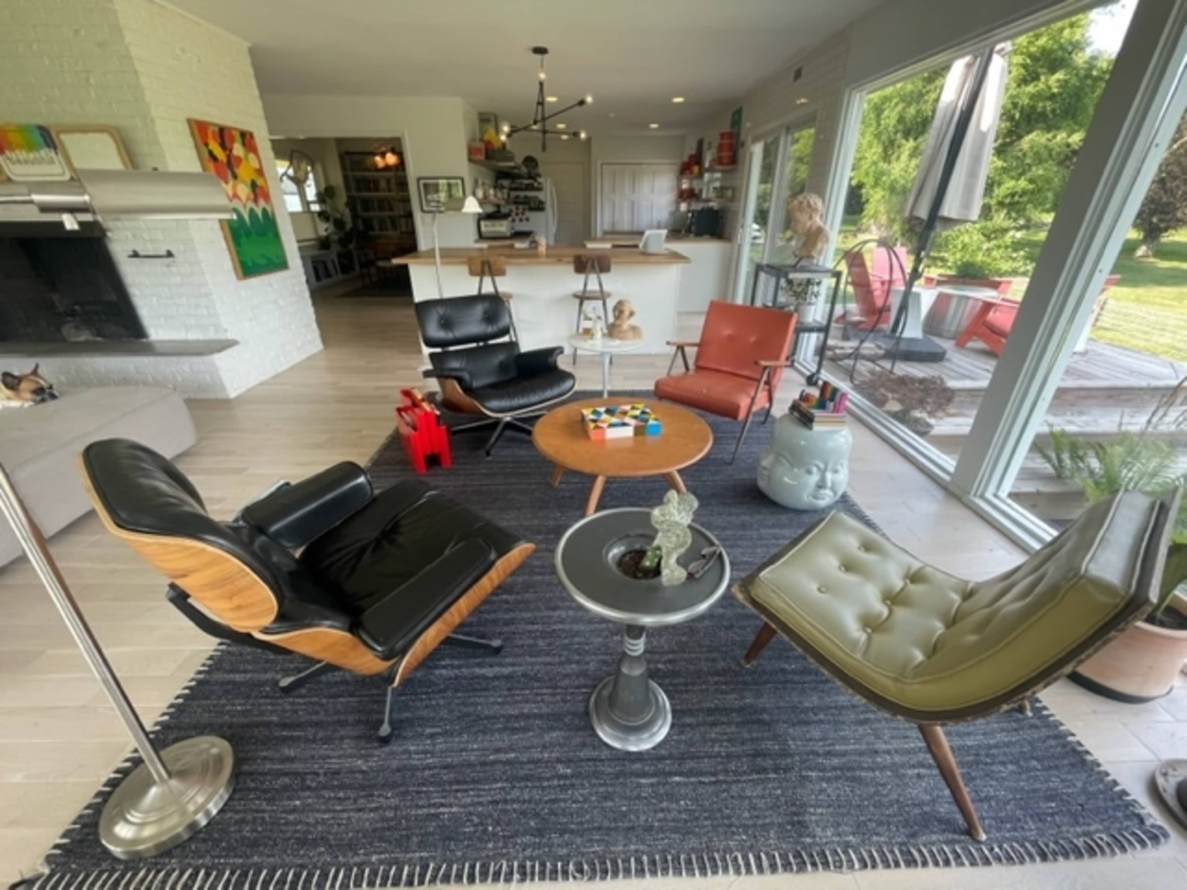 A modern living room features a mix of vintage furniture, including an Eames lounge chair, a coffee table with a colorful game, and large windows overlooking a garden.
