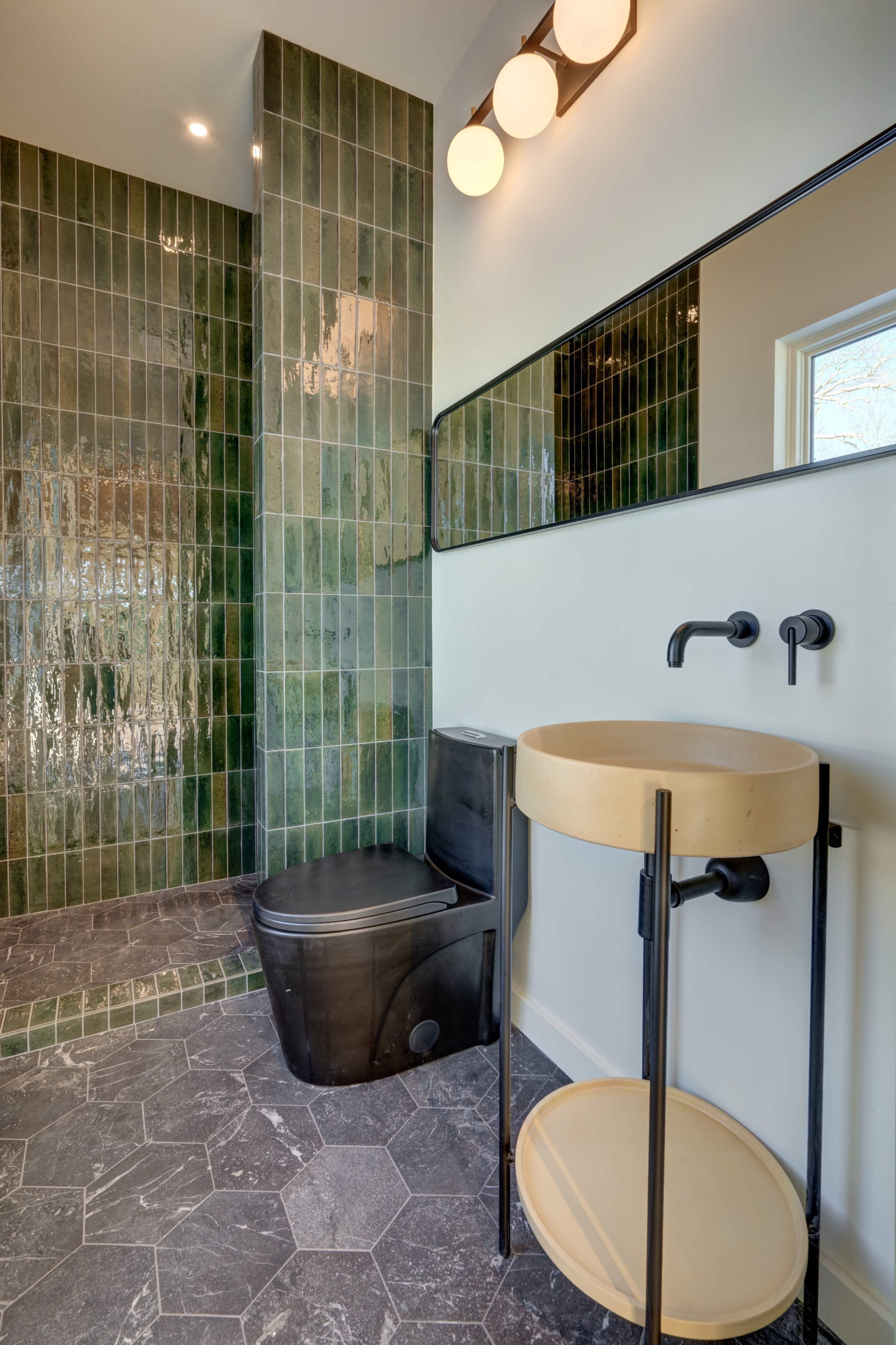 The image shows a modern bathroom with green tiled walls, a black toilet, a round bowl sink on a black metal stand, and a large mirror.