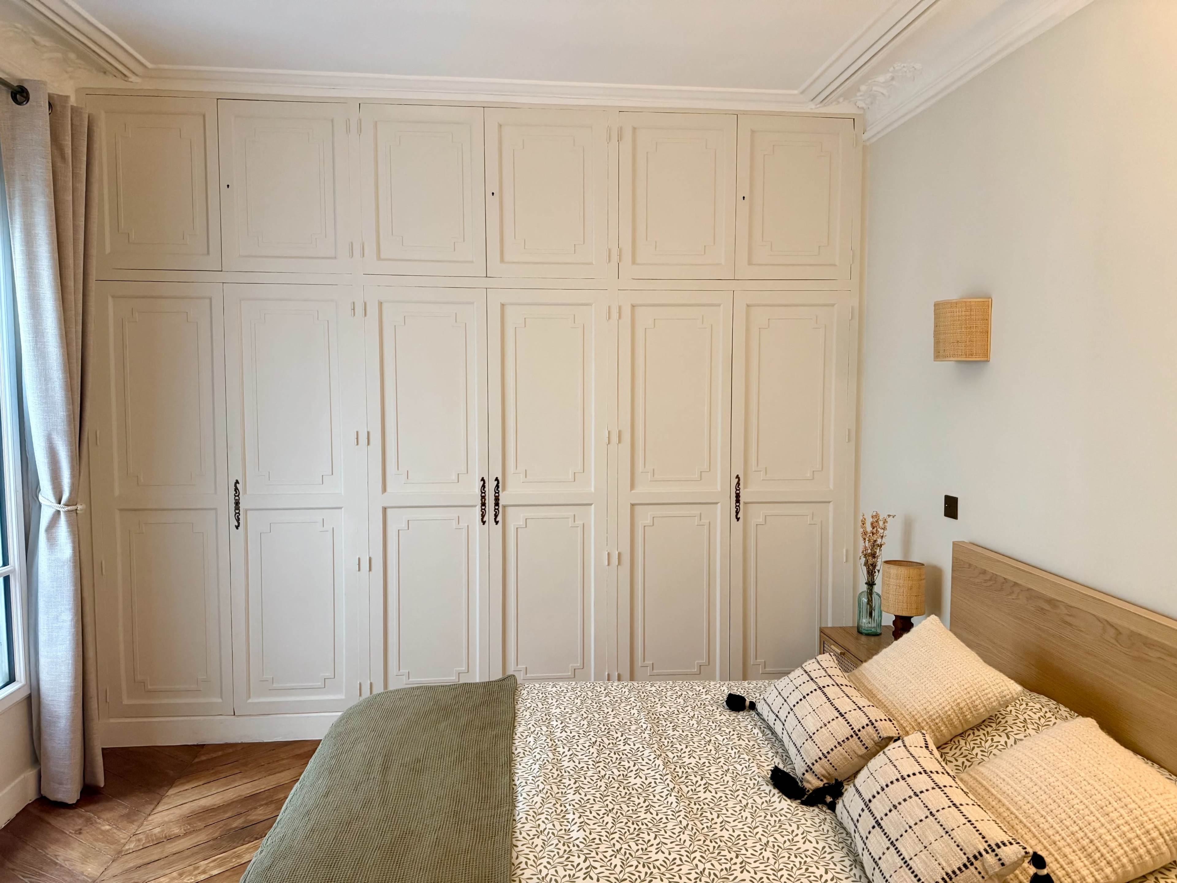 The image shows a well-organized bedroom with a wooden bed, patterned pillows, and a wall of closed cabinets.