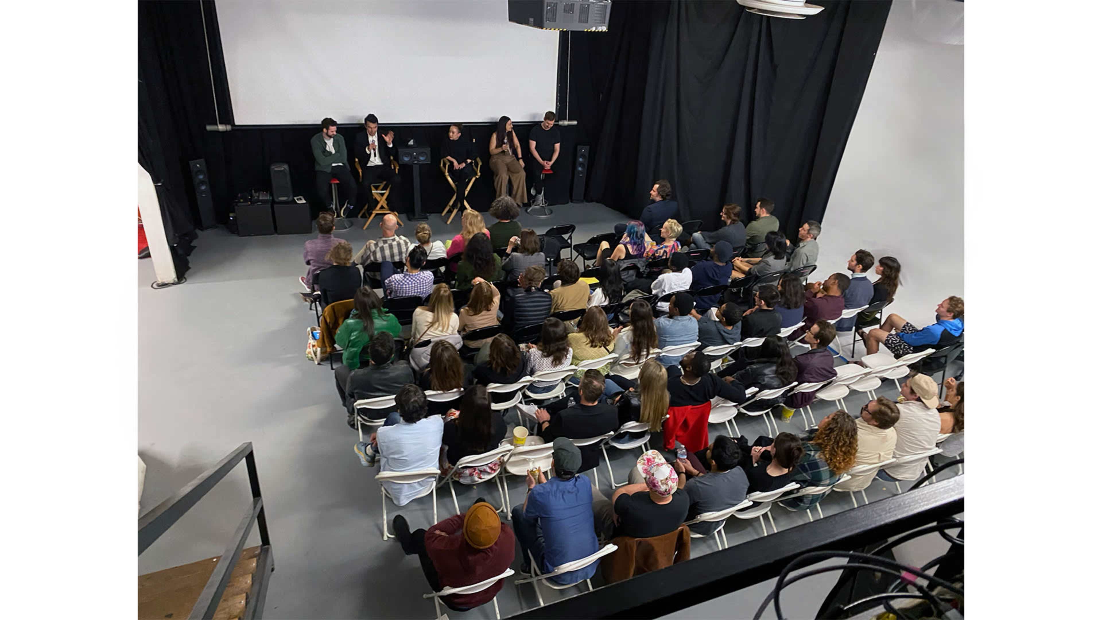 A large audience is seated in a theater setting, watching a panel discussion on a stage with multiple speakers.