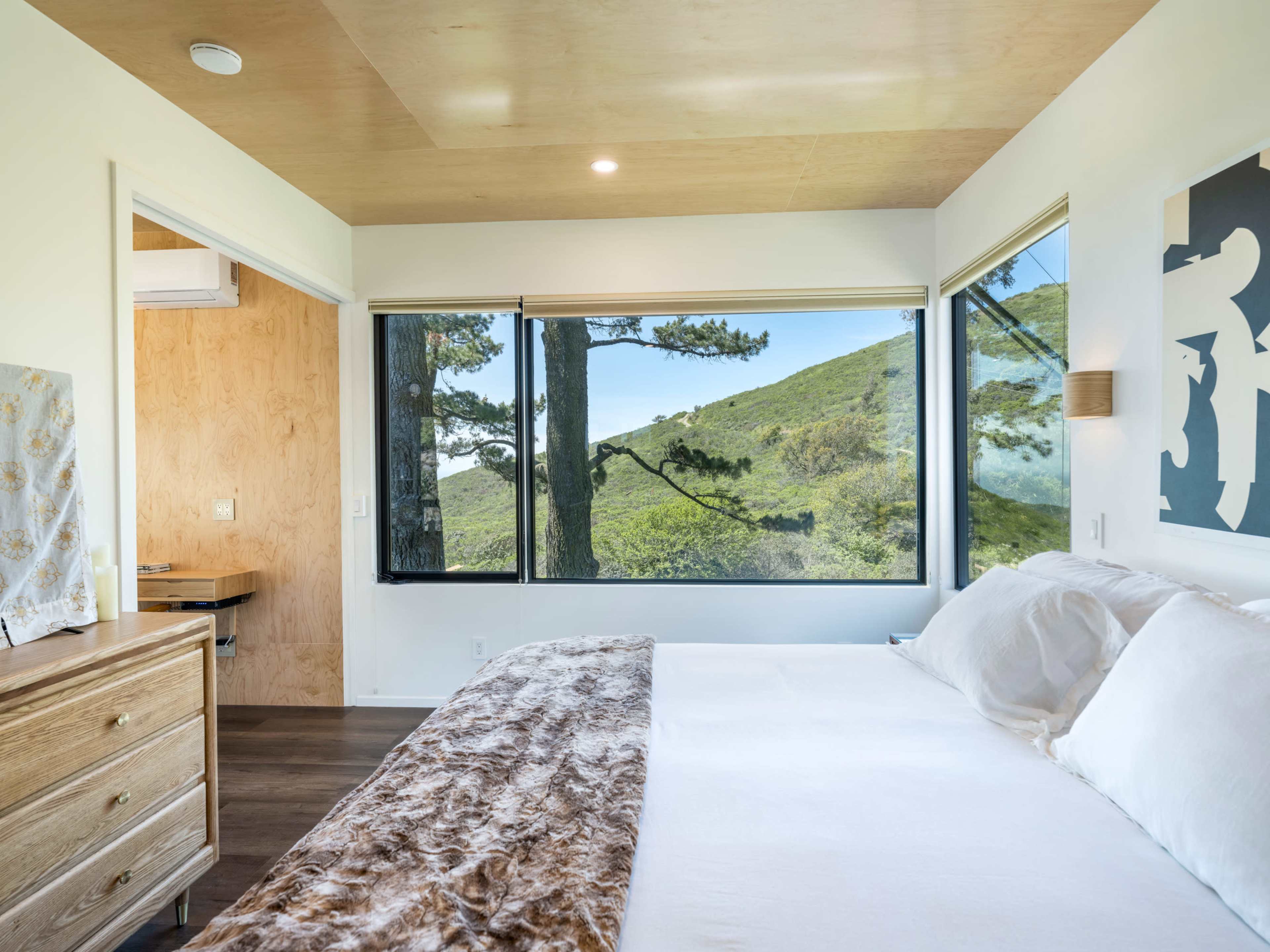 A modern bedroom features a bed with a fur throw and large windows overlooking a green hillside.