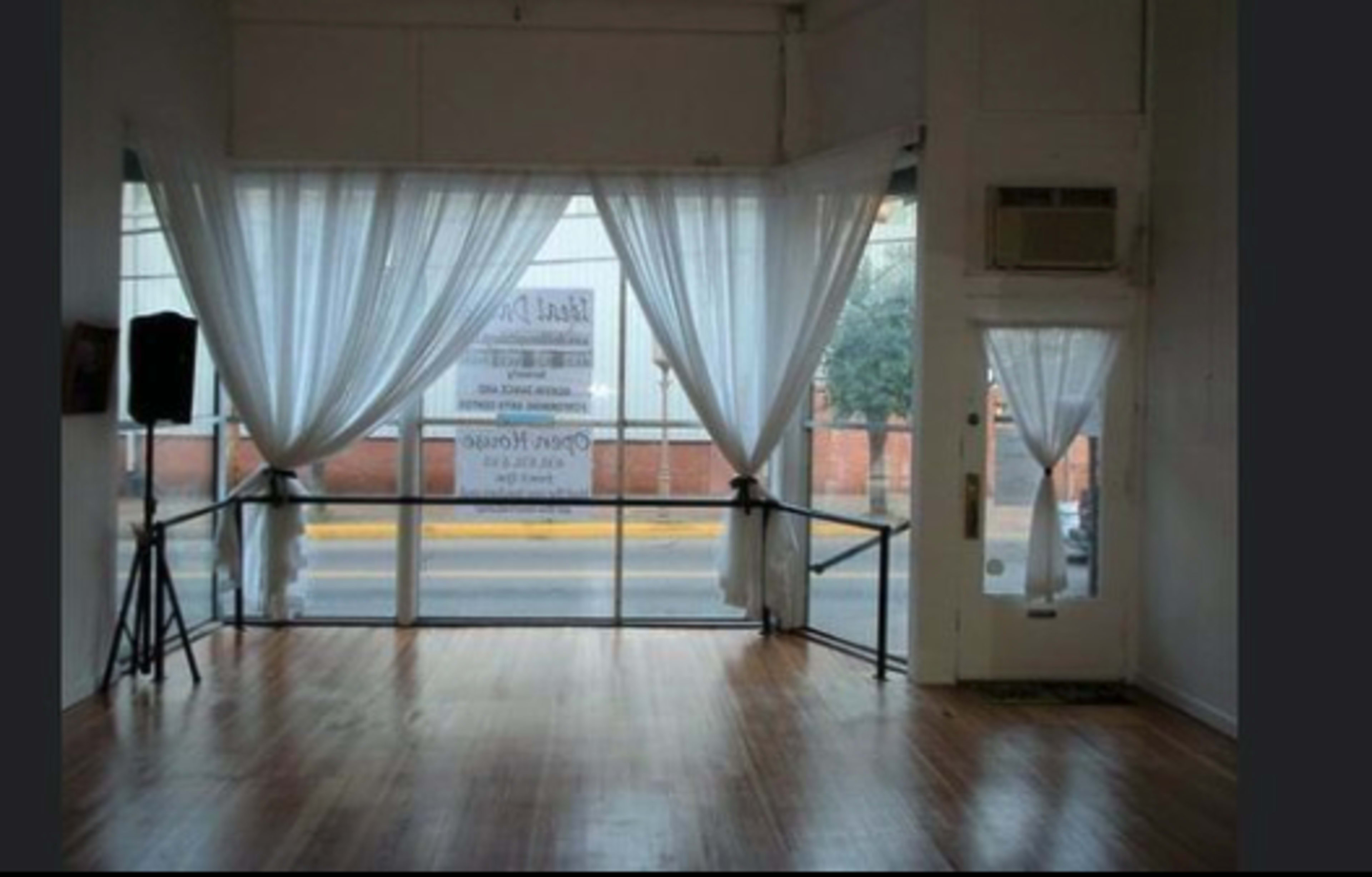 A spacious room with wooden flooring, large windows dressed in white curtains, and a view of a street outside.