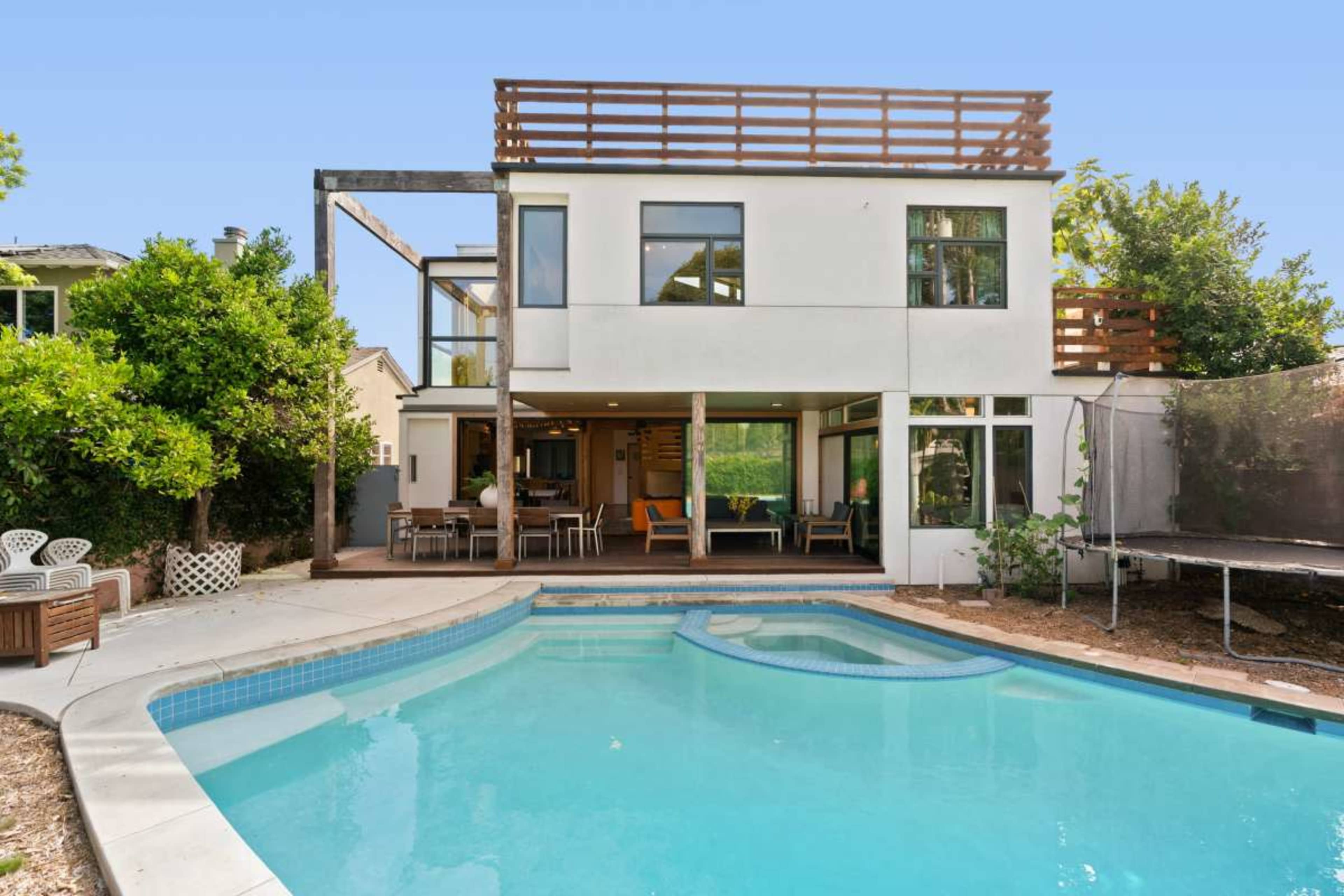 The image shows a modern two-story house with a pool in the backyard, featuring a spacious deck and a trampoline.