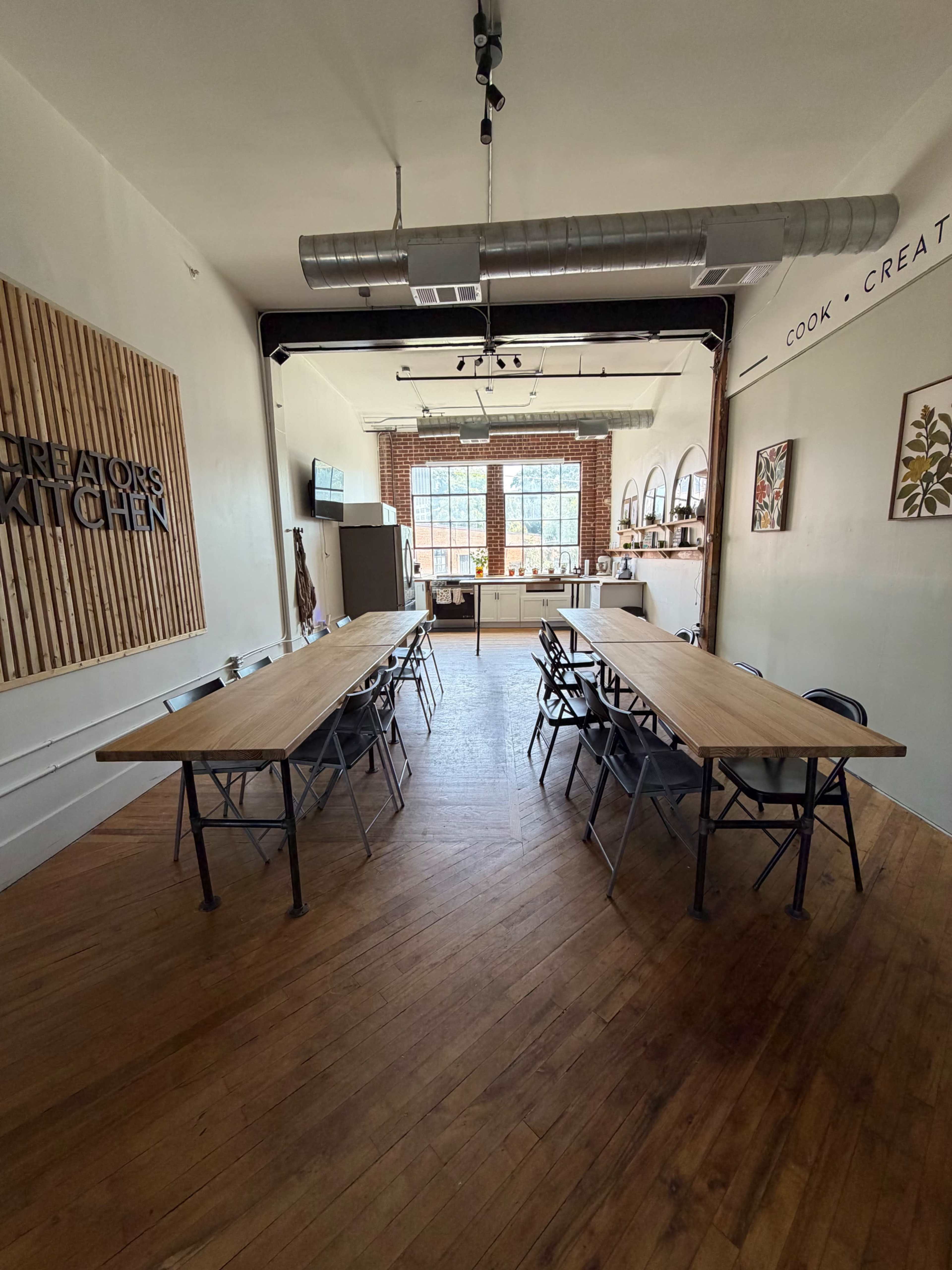 The image shows a spacious kitchen area with two long wooden tables set up for a gathering, illuminated by natural light from large windows.