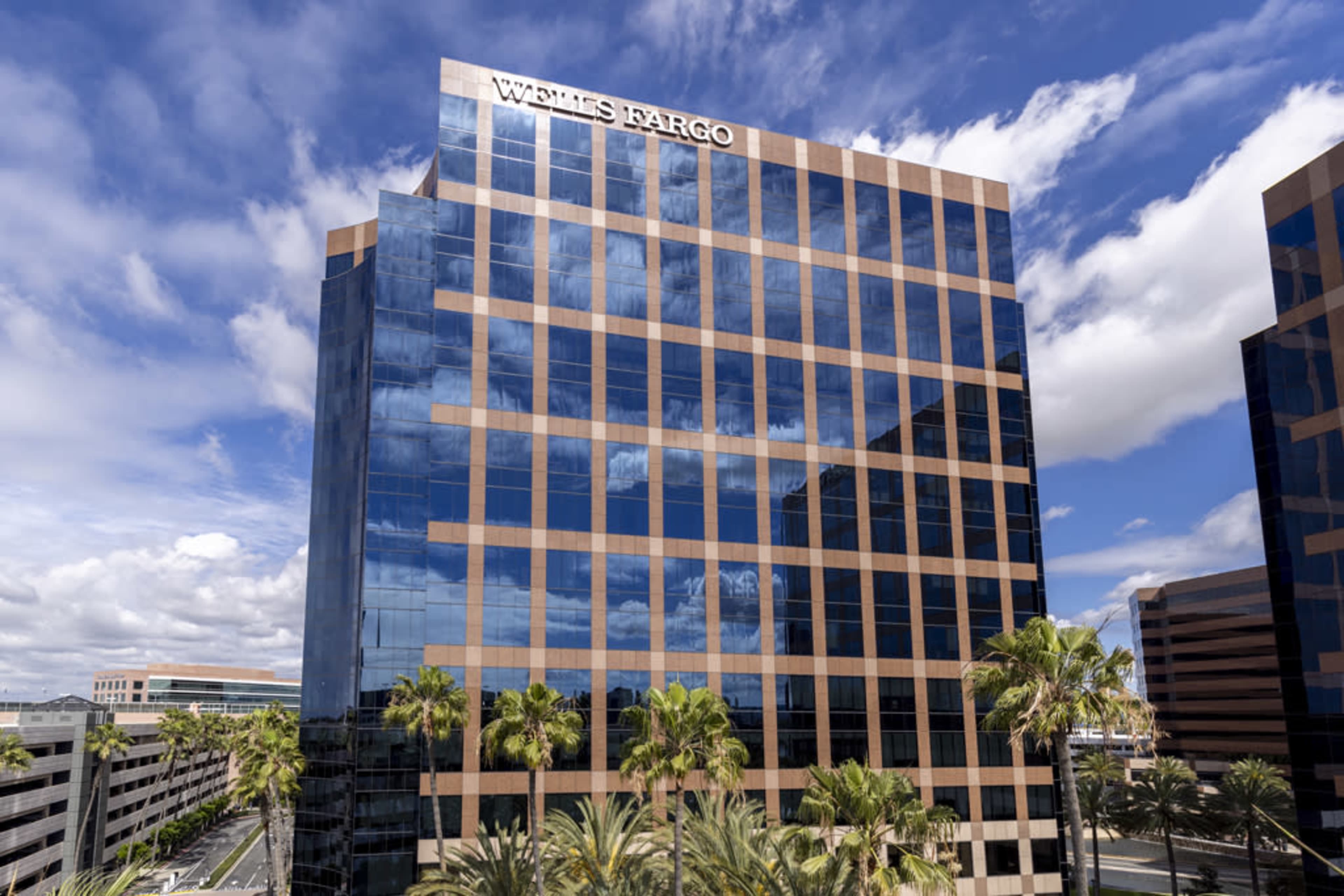 A tall office building with reflective glass windows displays the Wells Fargo logo at the top, set against a backdrop of blue sky and scattered clouds.
