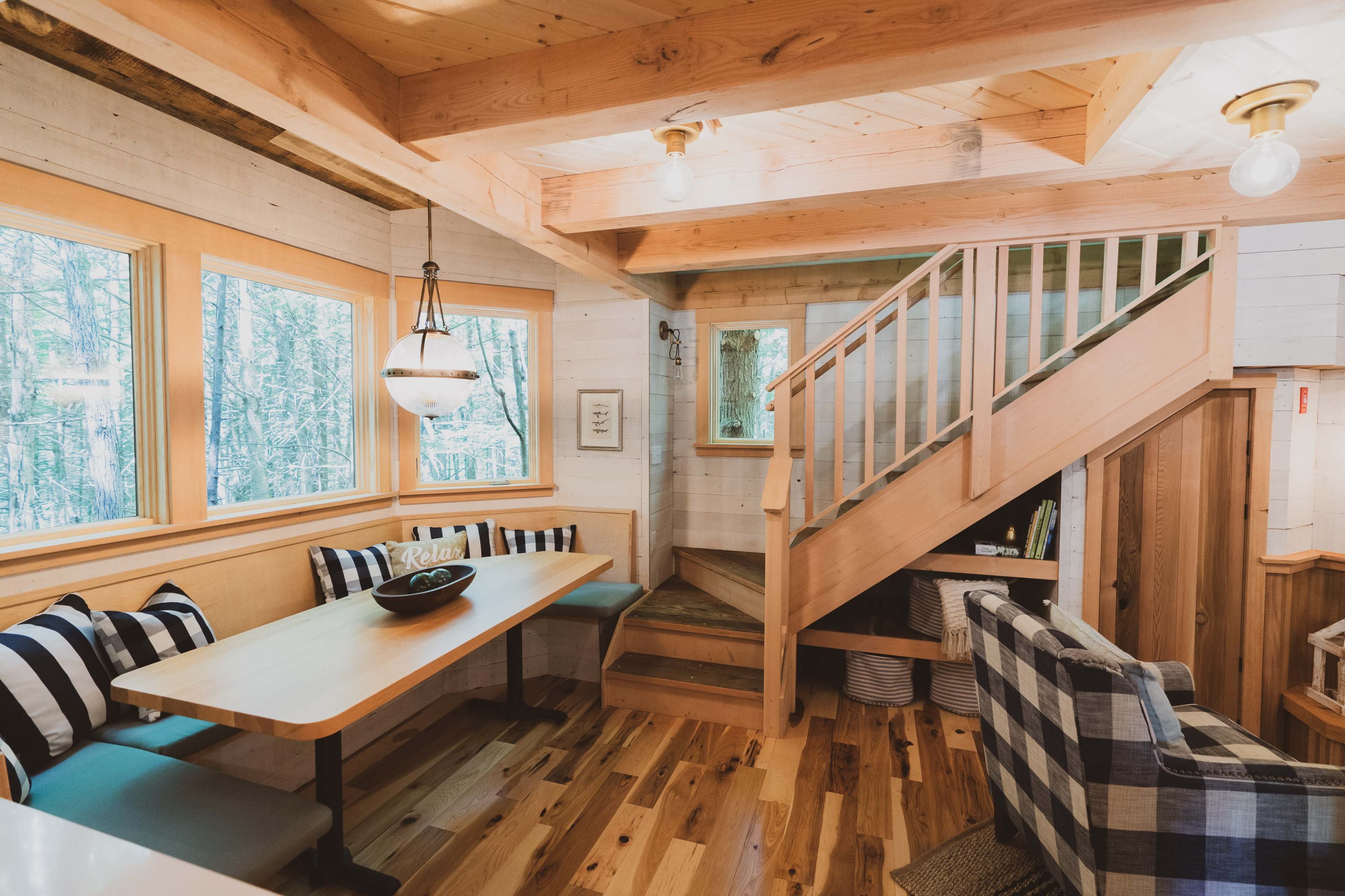 A cozy wooden interior with a dining area featuring a table and bench, a staircase, and large windows that allow natural light to fill the space.