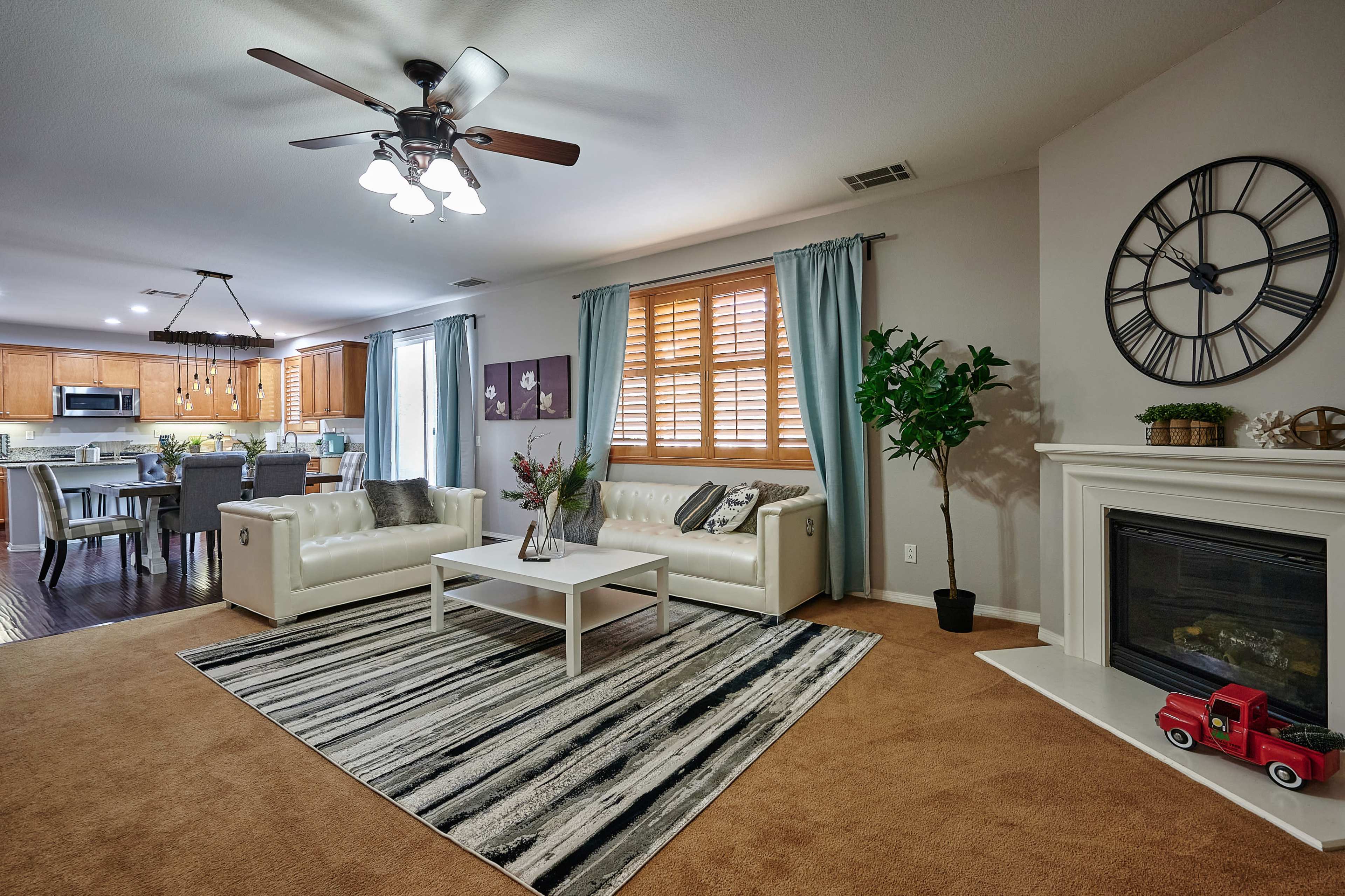 A living room with white leather sofas, a coffee table on a striped rug, a fireplace, and a kitchen visible in the background.
