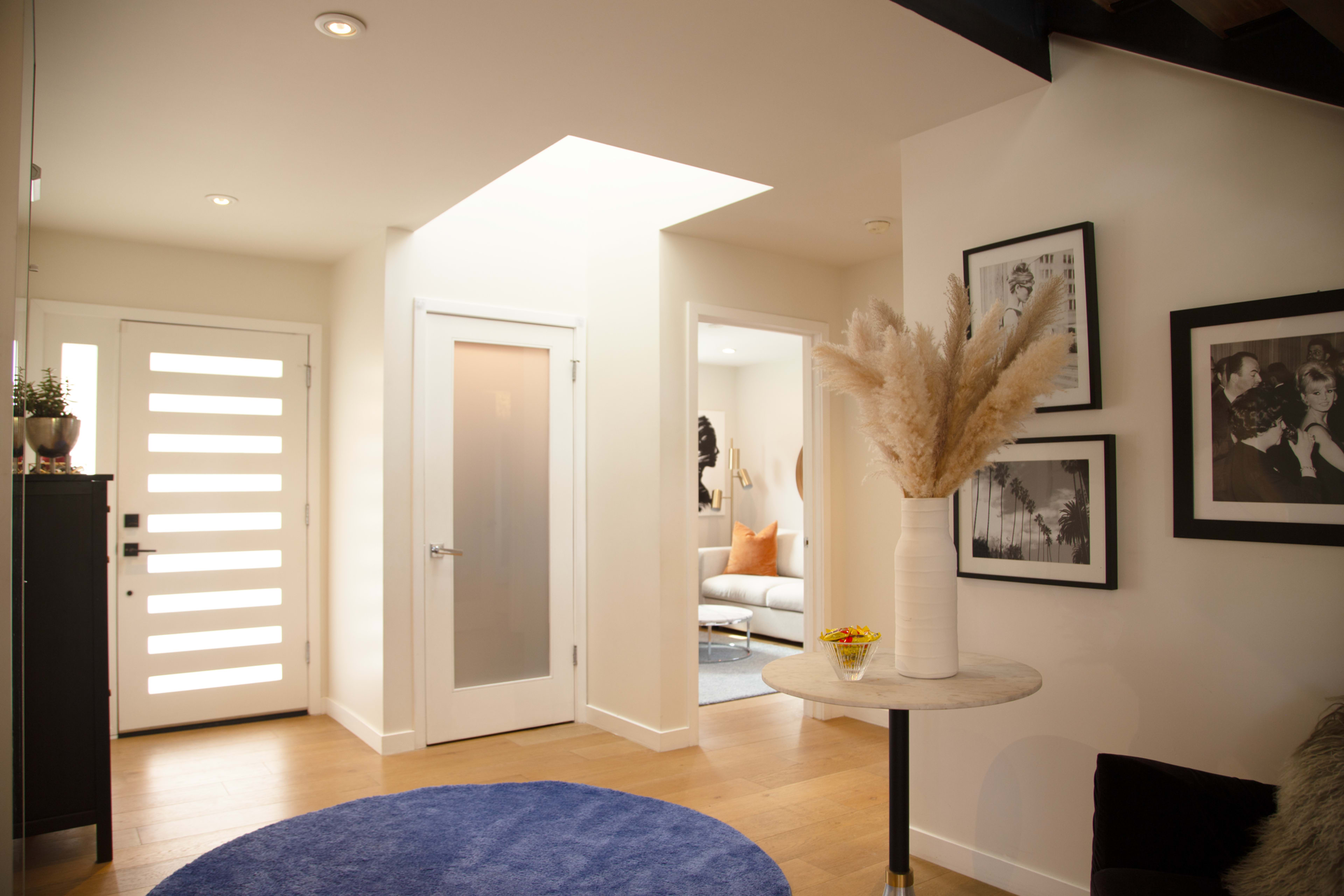 The image shows a modern hallway with a white entrance door, a small circular table with a vase, and framed photographs on the wall.