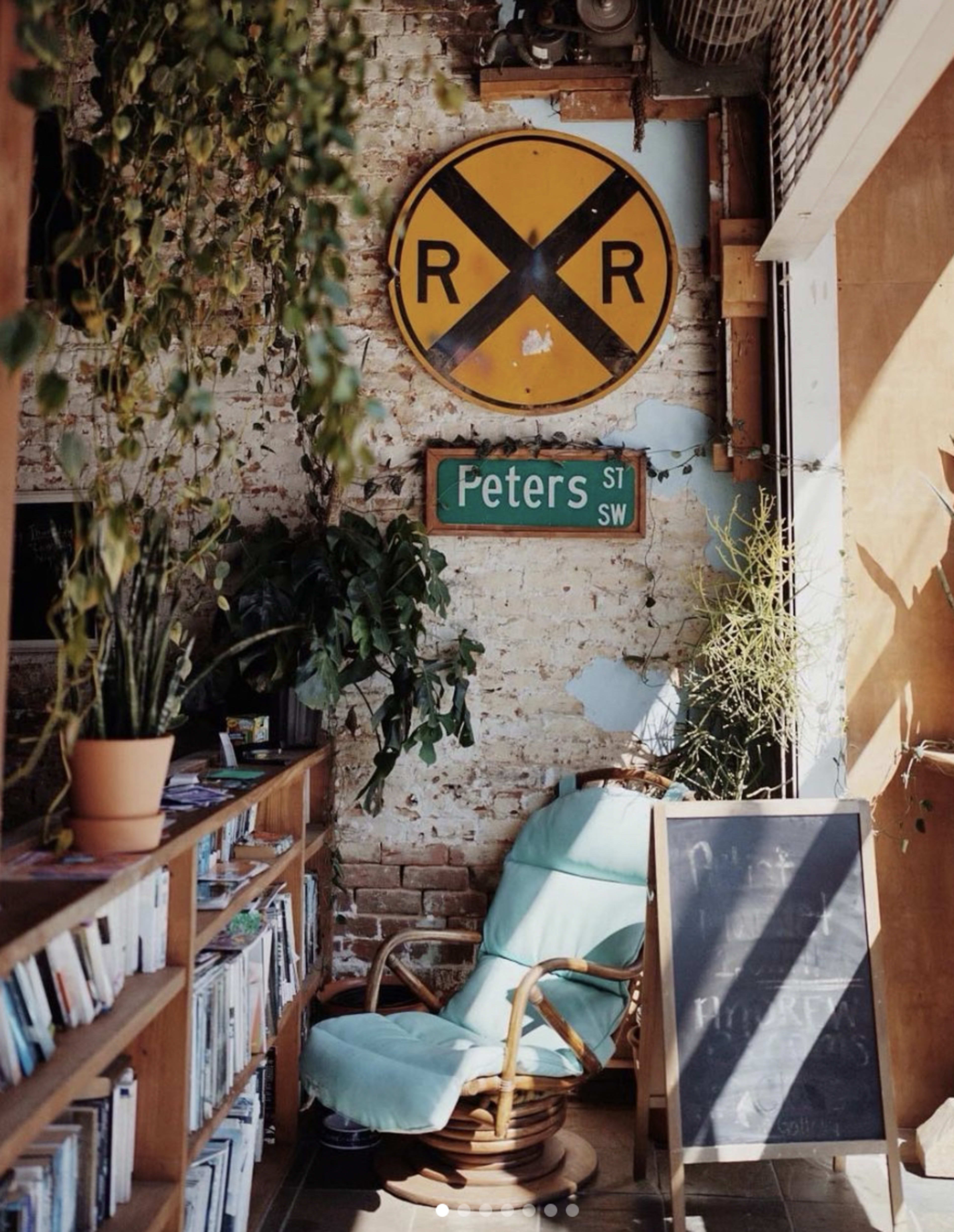 A cozy reading nook with a rocking chair beside a bookshelf, adorned with plants and vintage signs on a rustic wall.