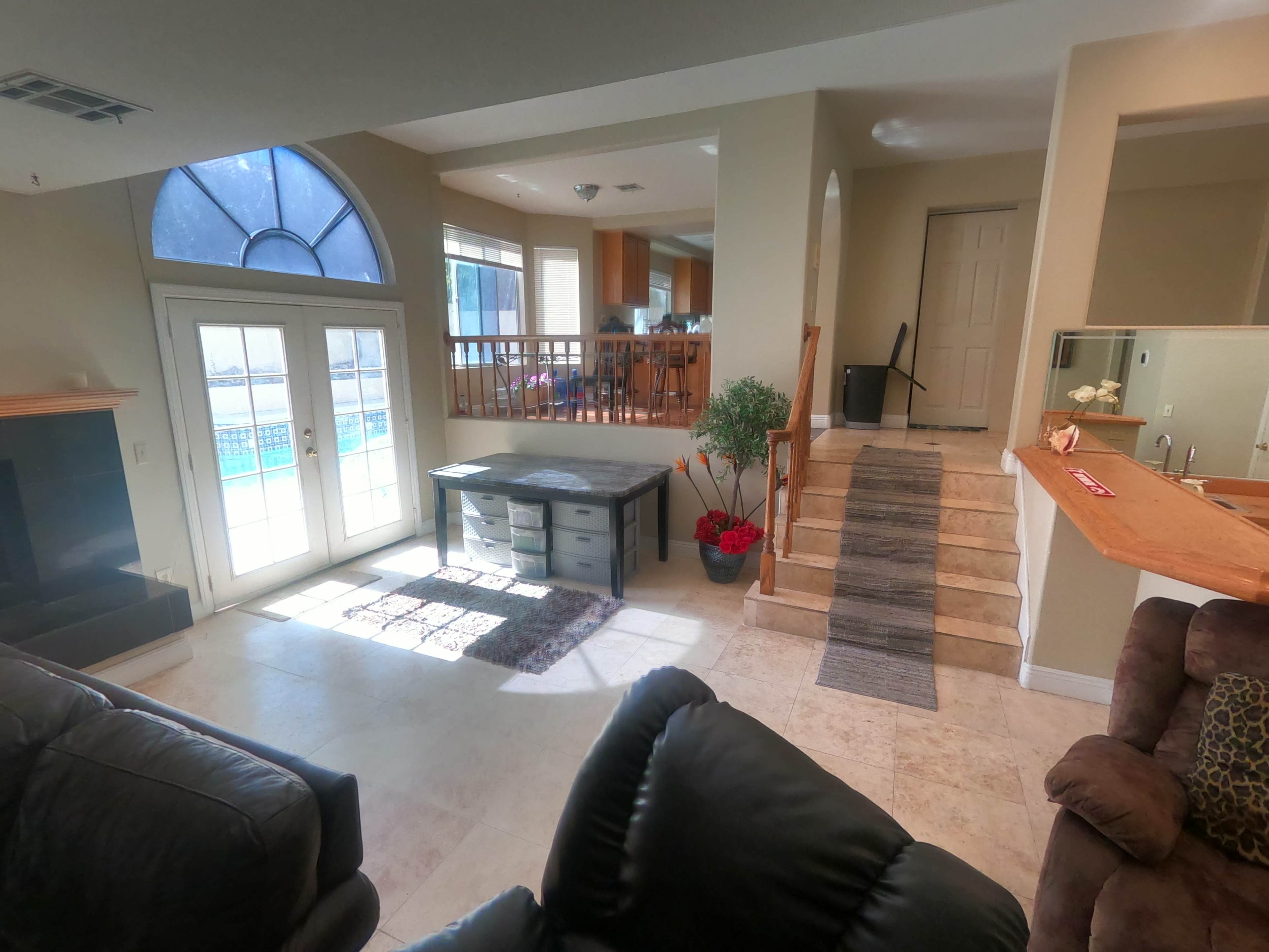 The image shows a spacious living area with a large window, a staircase leading to another level, and a view of a pool area through the glass doors.