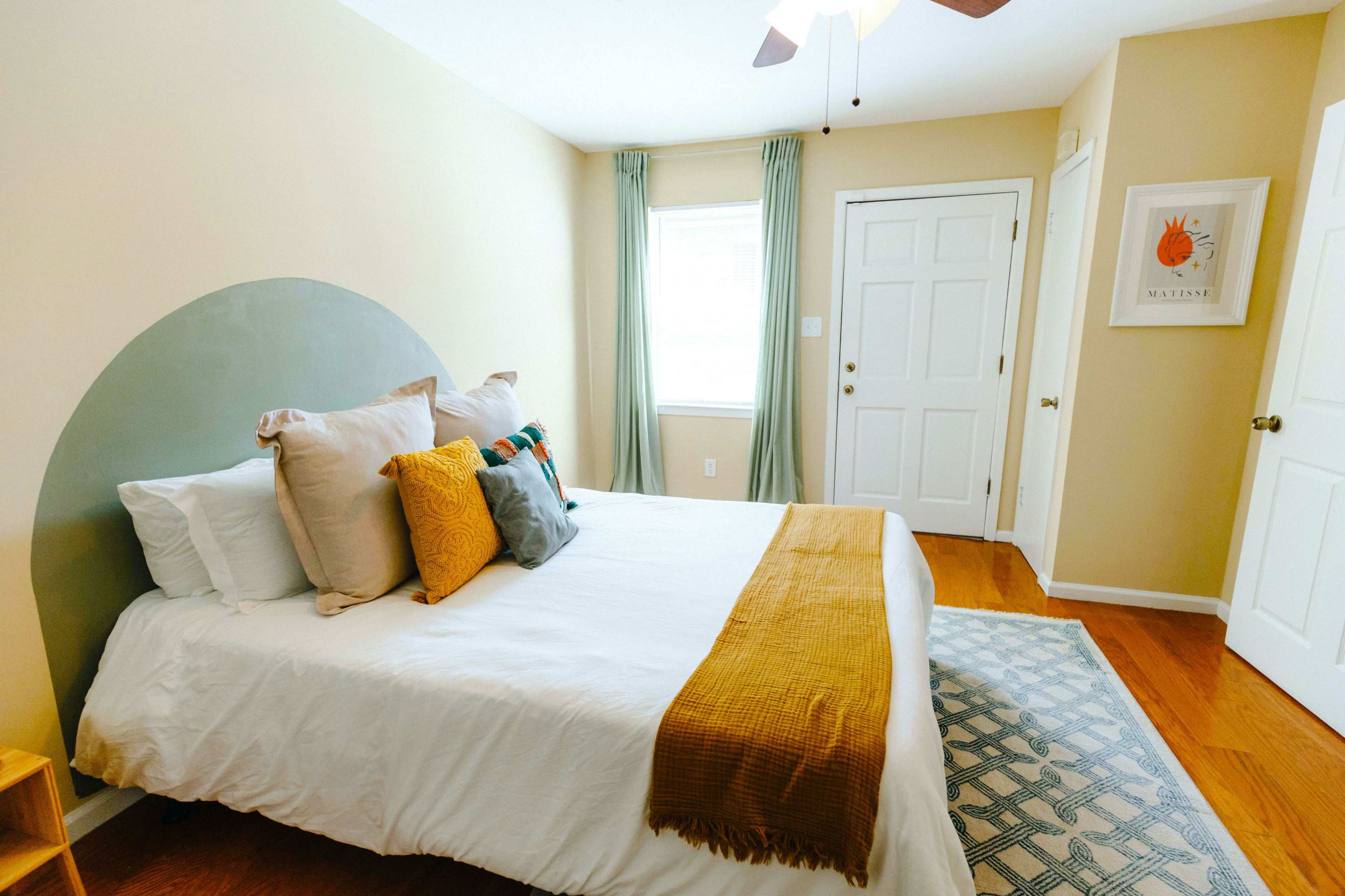 A brightly lit bedroom featuring a bed with multiple pillows, a mustard-colored throw blanket, and a framed artwork on the wall.