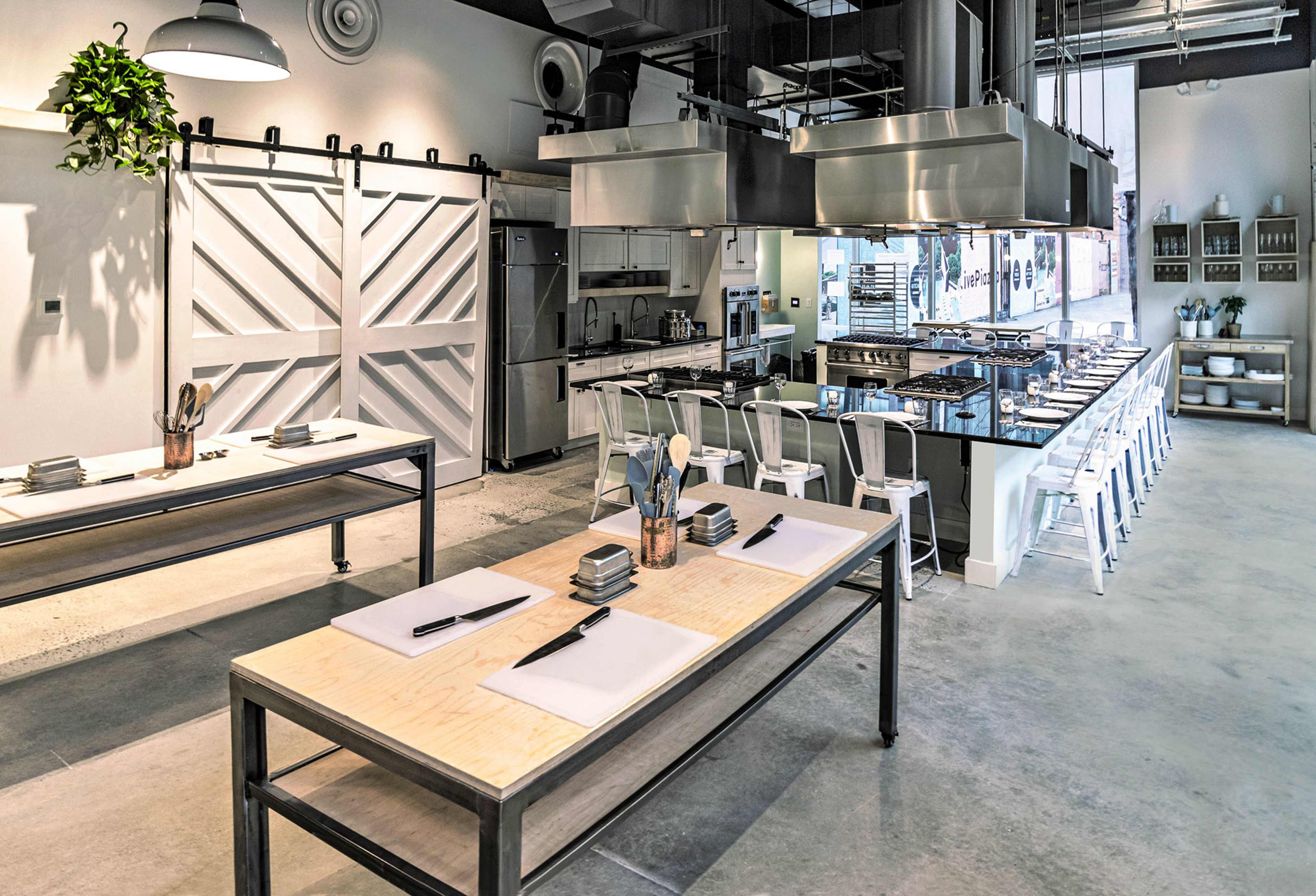 A modern kitchen workspace features metal tables, kitchen equipment, and a row of stools against a minimalist design backdrop.