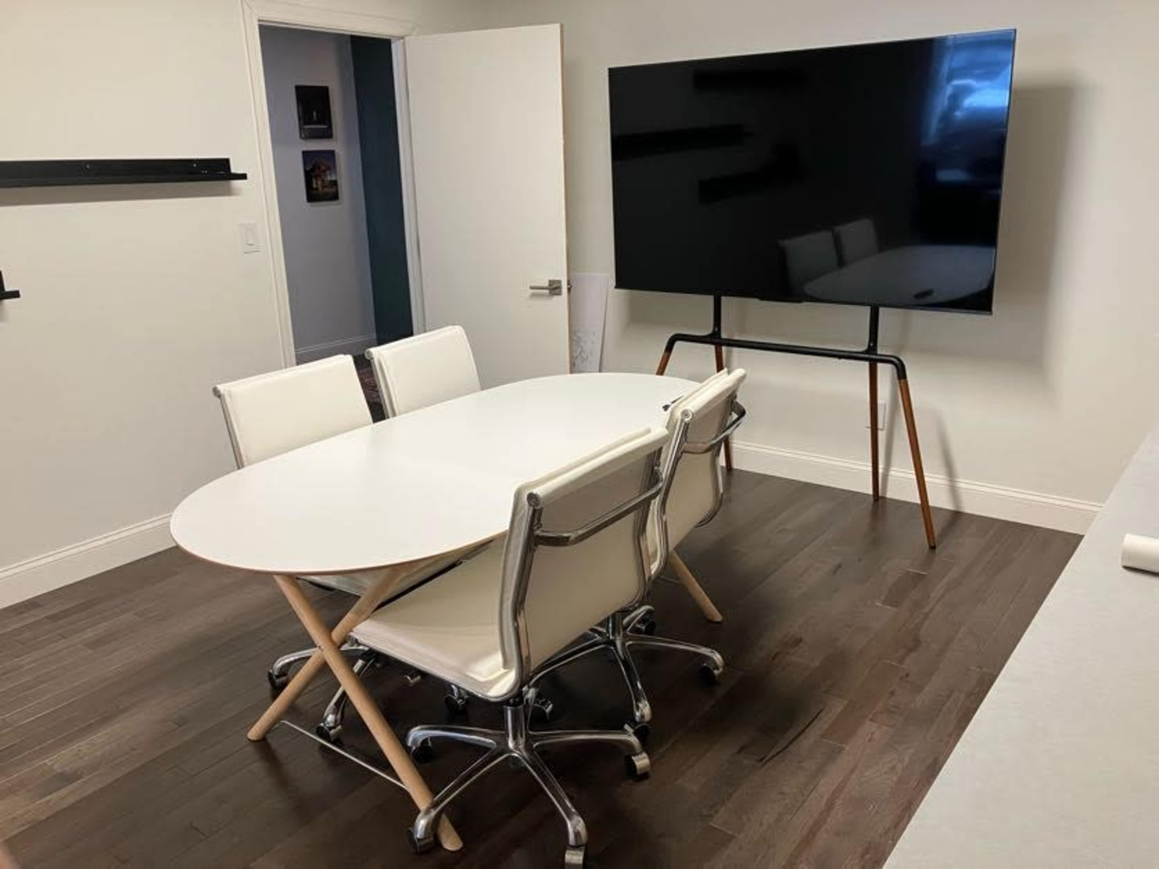 A conference room features a white oval table surrounded by four chrome office chairs and a large television mounted on a stand.
