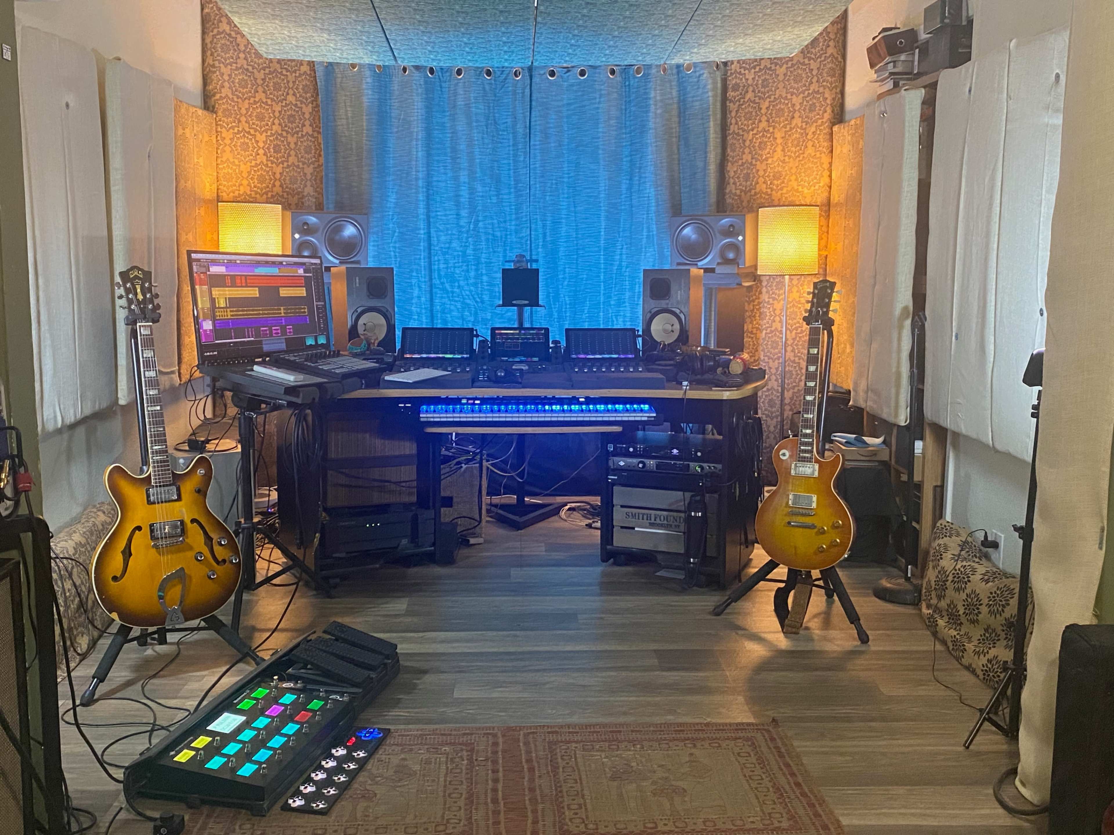 A music studio setup featuring two electric guitars, multiple monitors, audio equipment, and a MIDI controller, all arranged in a cozy room with soundproofing panels.