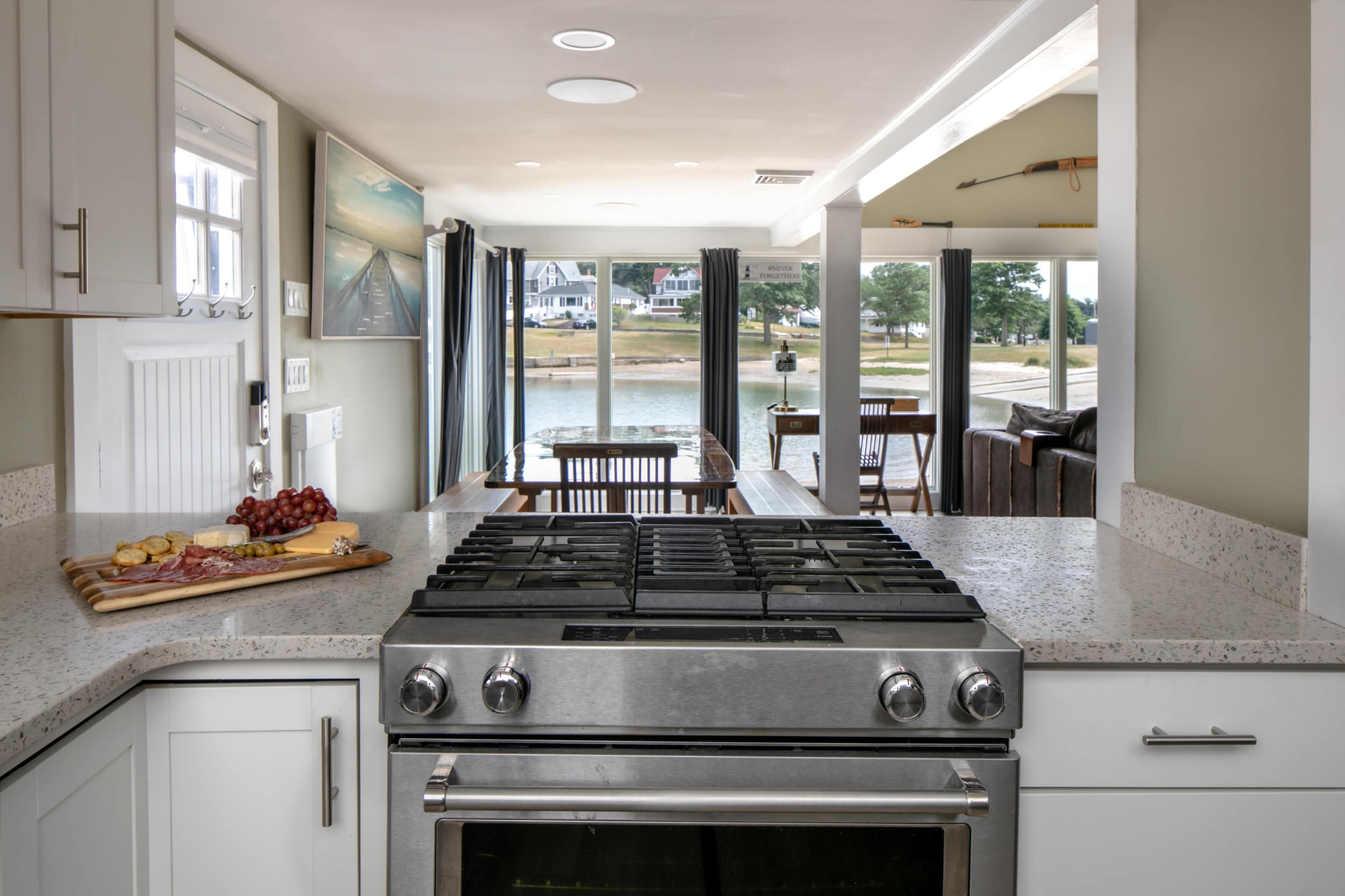 A modern kitchen with a stainless steel stove overlooks a dining area and waterfront through large windows.