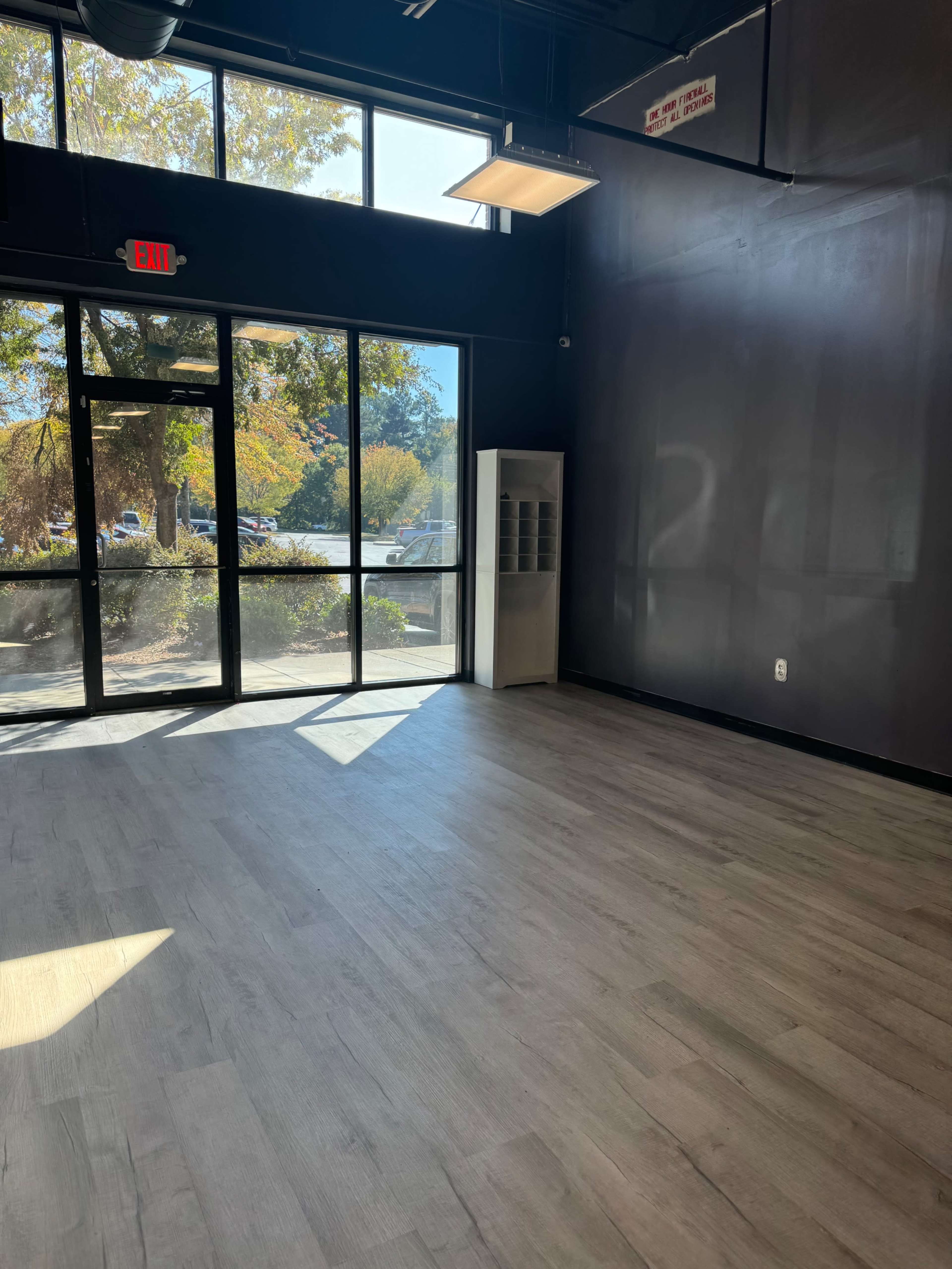 The image shows a spacious, empty room with large windows allowing natural light to enter and a door leading outside.