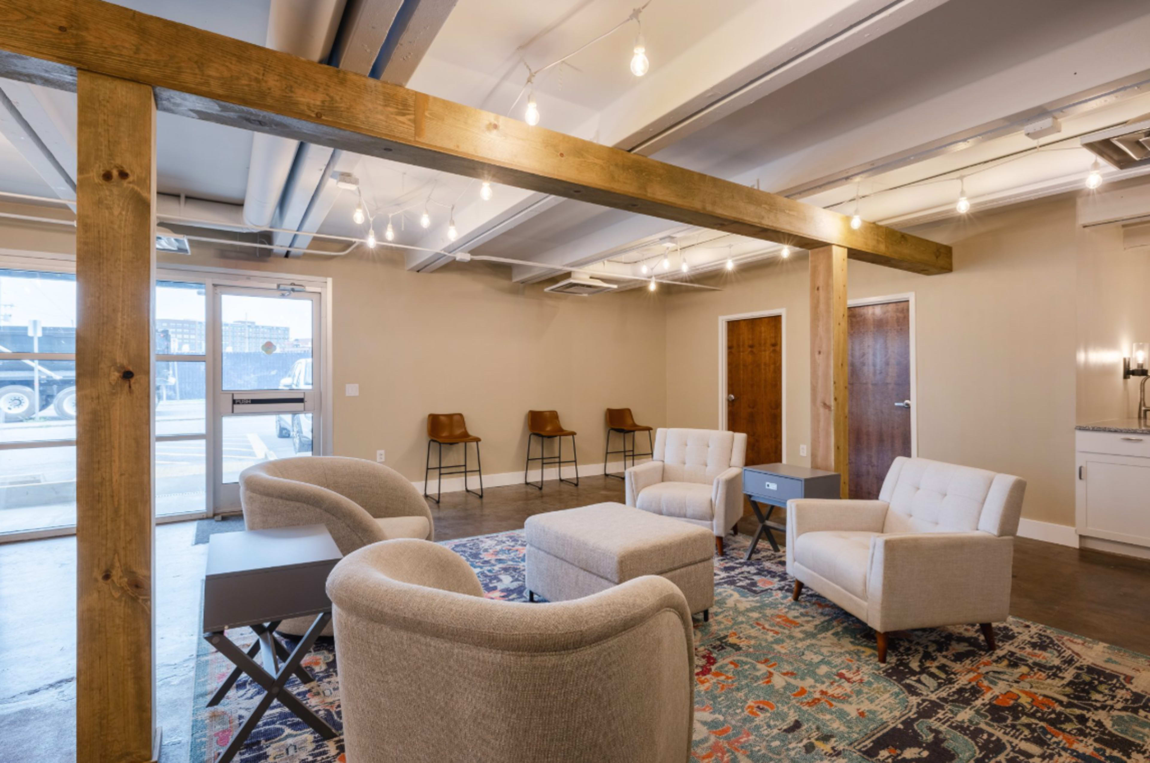 A modern, minimalist lounge area features four light-colored armchairs arranged around a patterned rug, with wooden beams and pendant lights enhancing the space's ambiance.