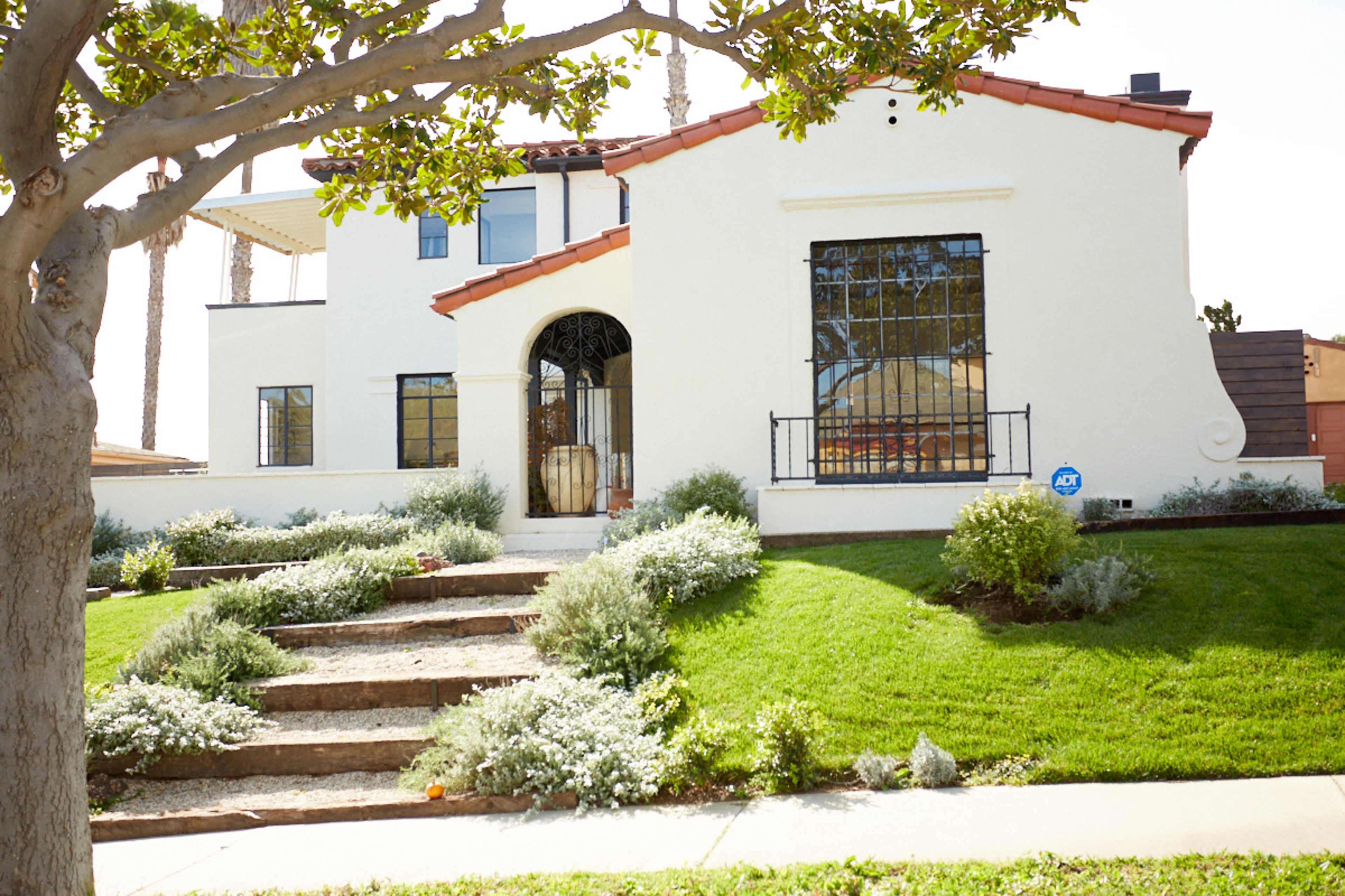 A two-story white house with a red-tiled roof is situated on a grassy lawn, featuring a stone walkway leading to the entrance.