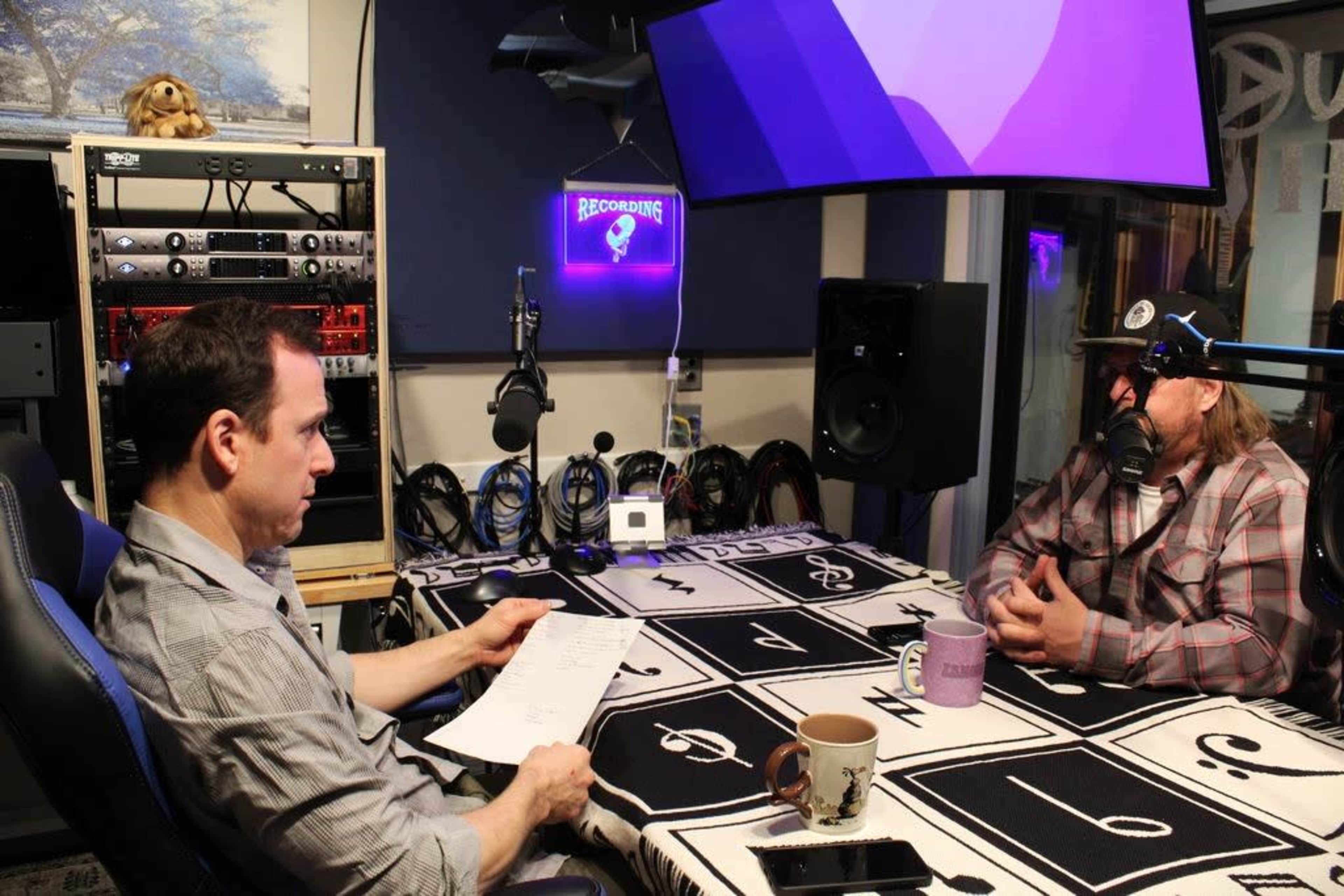 Two men are seated in a recording studio, one reading notes while the other listens, with audio equipment and a purple-lit screen in the background.