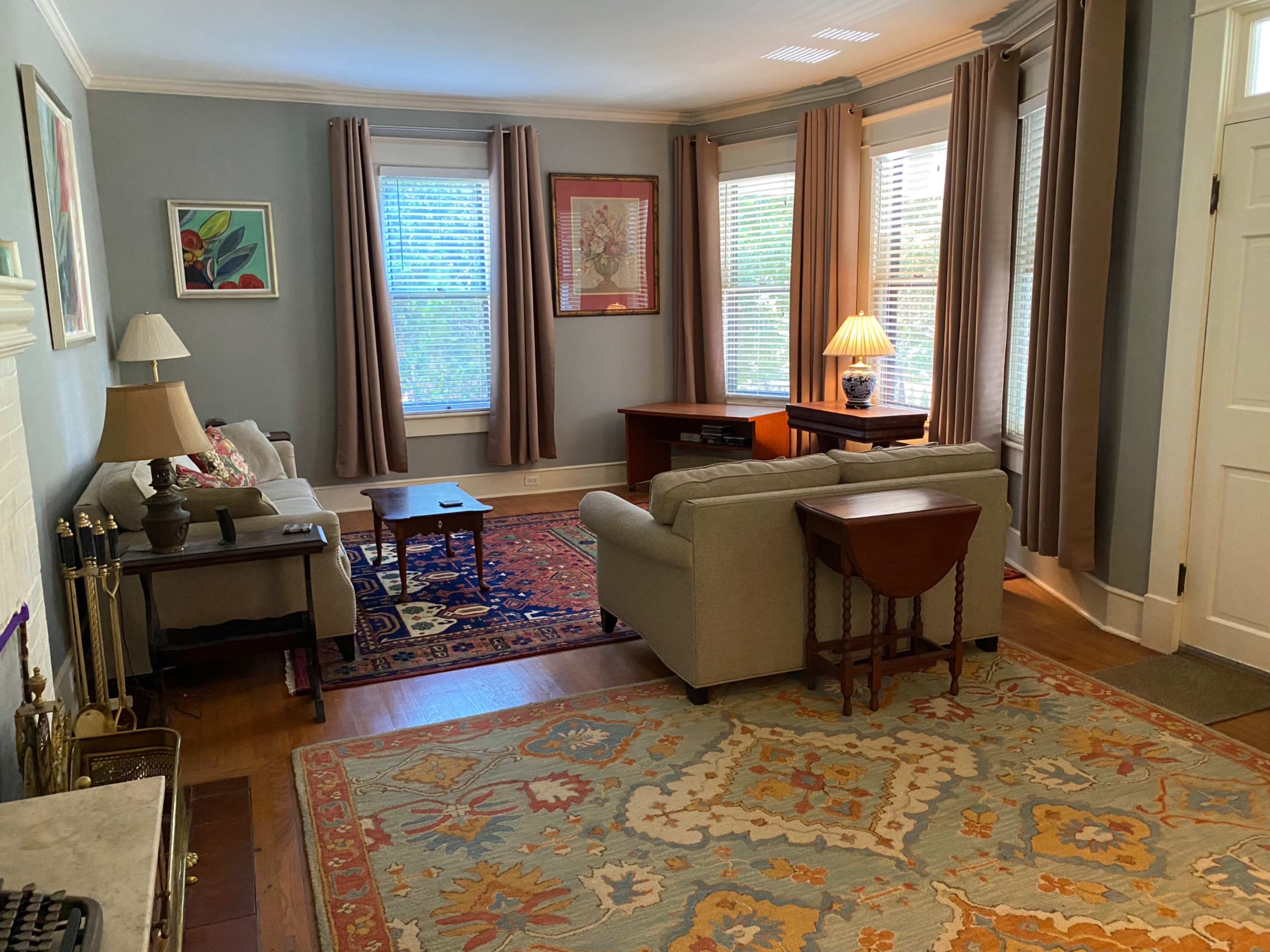 The image shows a living room with a sofa, armchair, side tables, and a large patterned rug, illuminated by natural light from the windows.