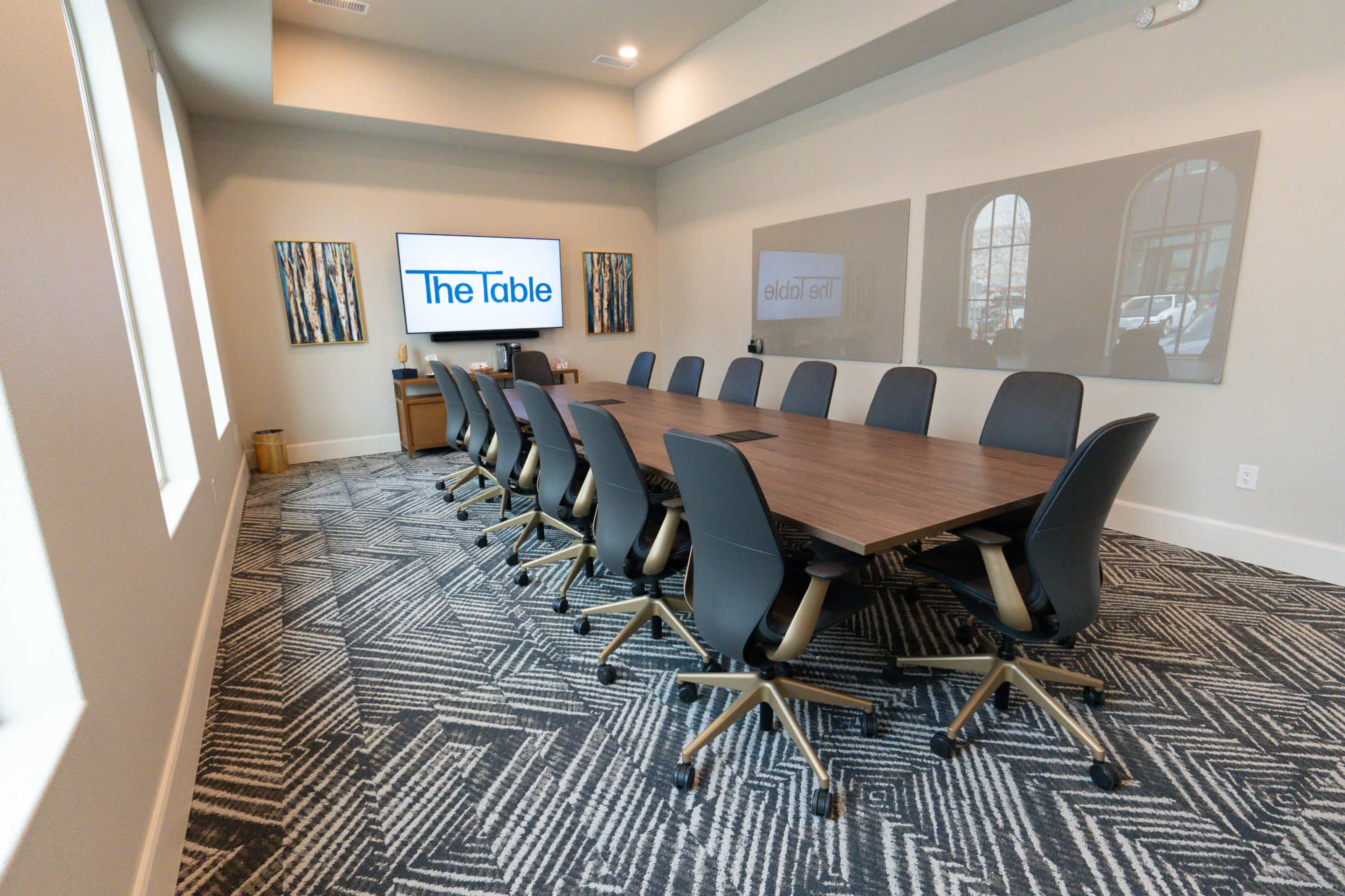 A conference room features a long wooden table surrounded by ergonomic black chairs, with a large screen displaying "The Table" on one wall and two framed artworks on another.