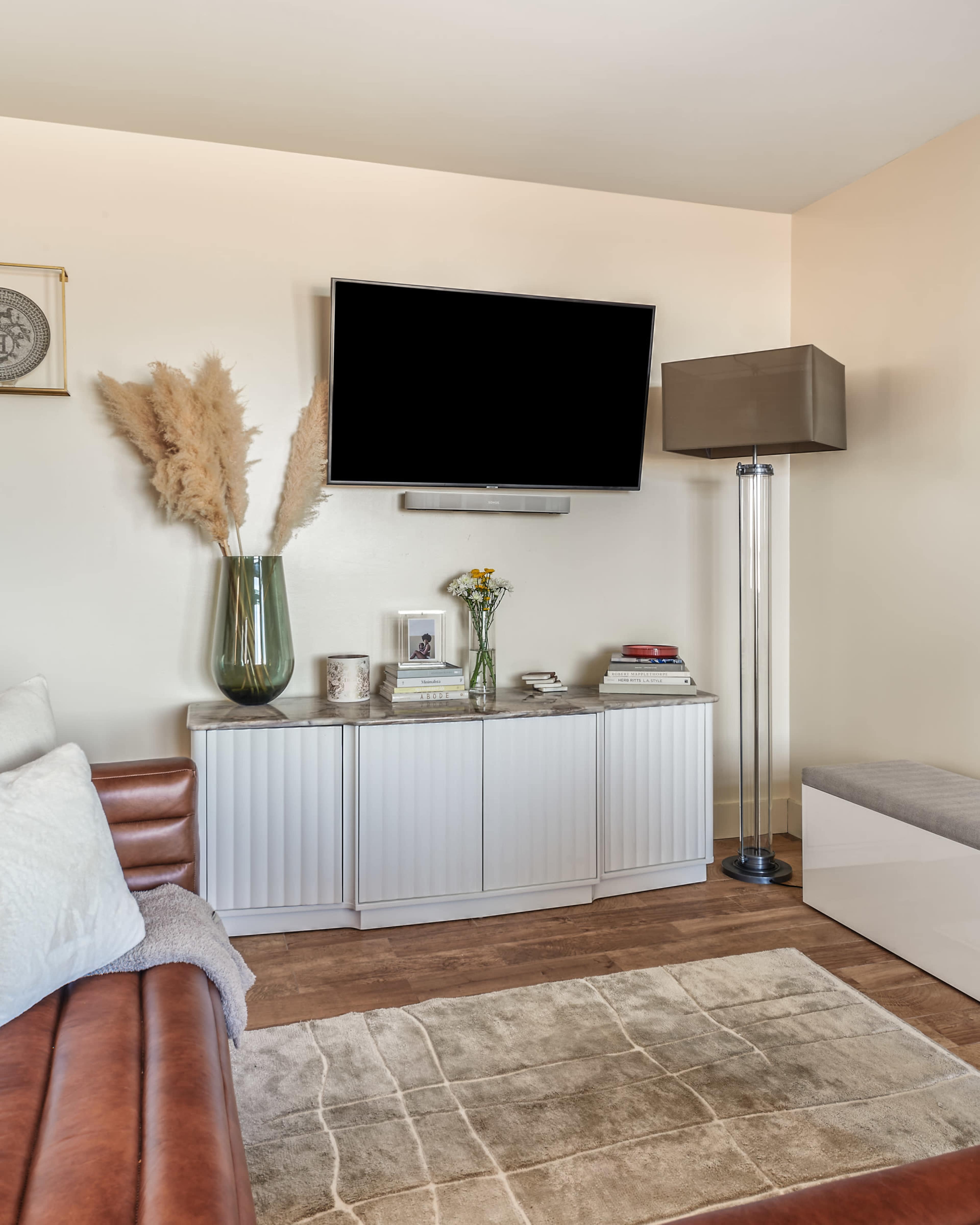 The scene features a living room with a mounted television above a white cabinet, a tall lamp, and decorative items including a vase with dried flowers.