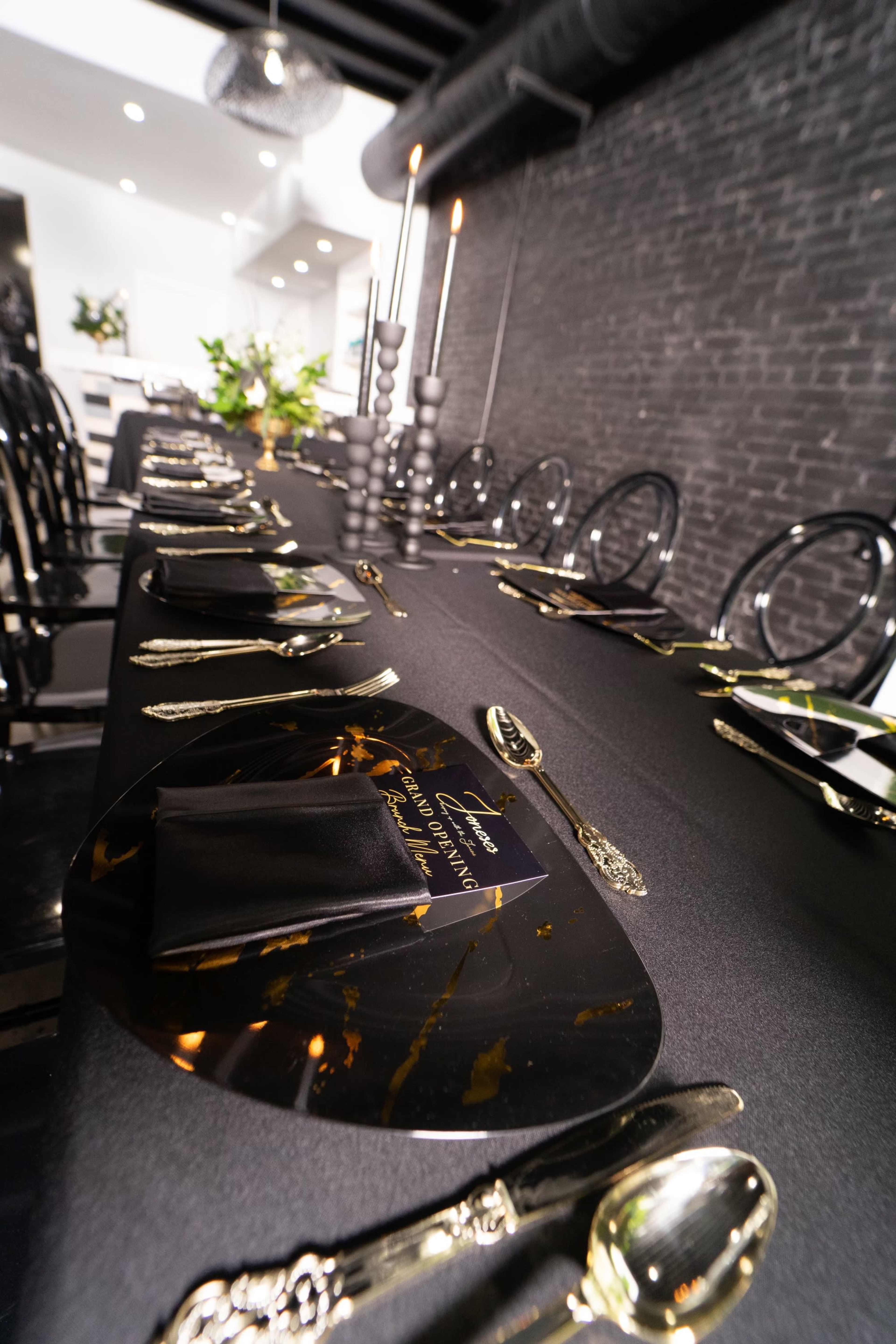 A long dining table is set with black and gold tableware, black napkins, and decorative candles, against a backdrop of dark brick walls.