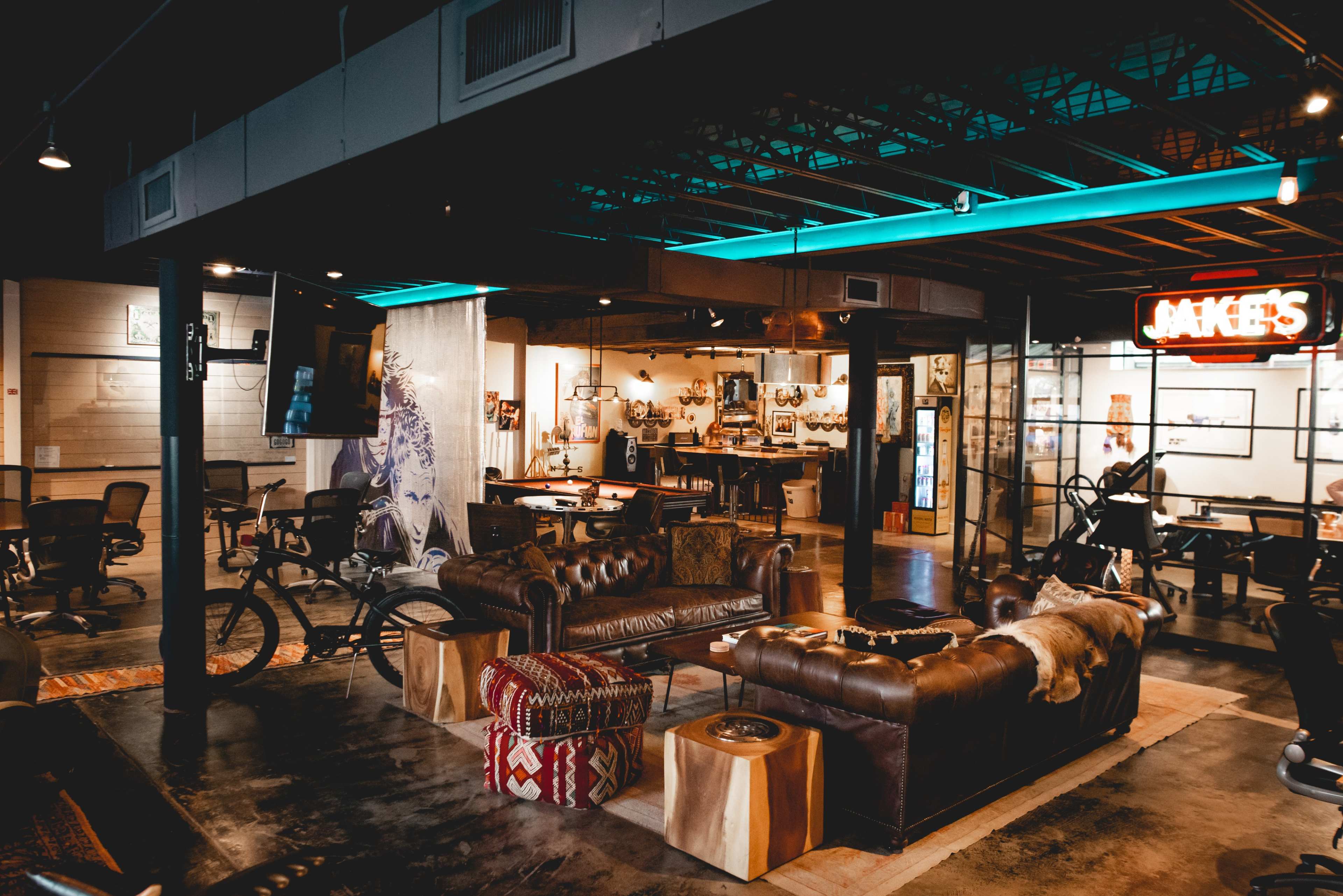The image depicts a modern office space featuring leather sofas, a wooden table, and a neon sign that reads "Jake's."