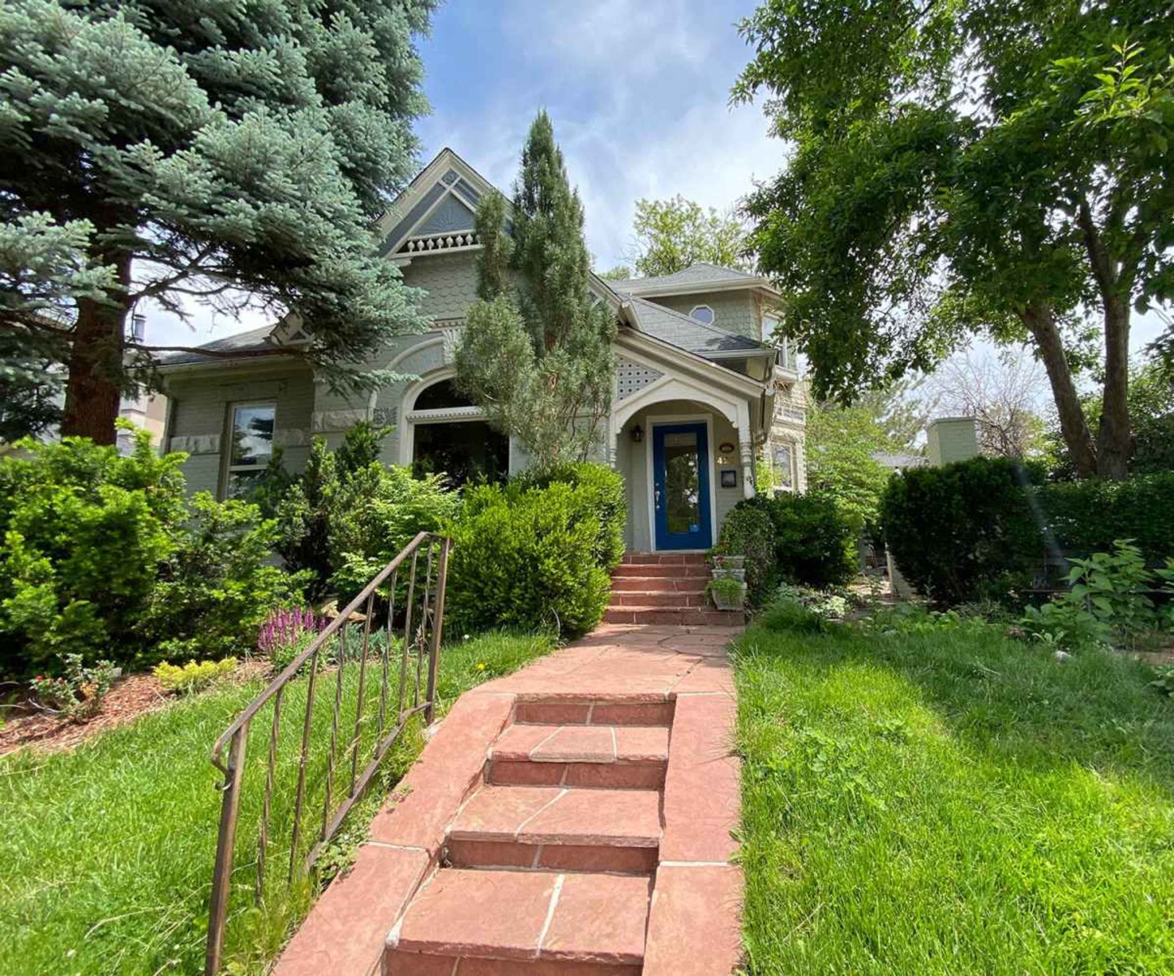 A vintage-style house with a blue front door, surrounded by lush greenery and stone steps leading to the entrance.