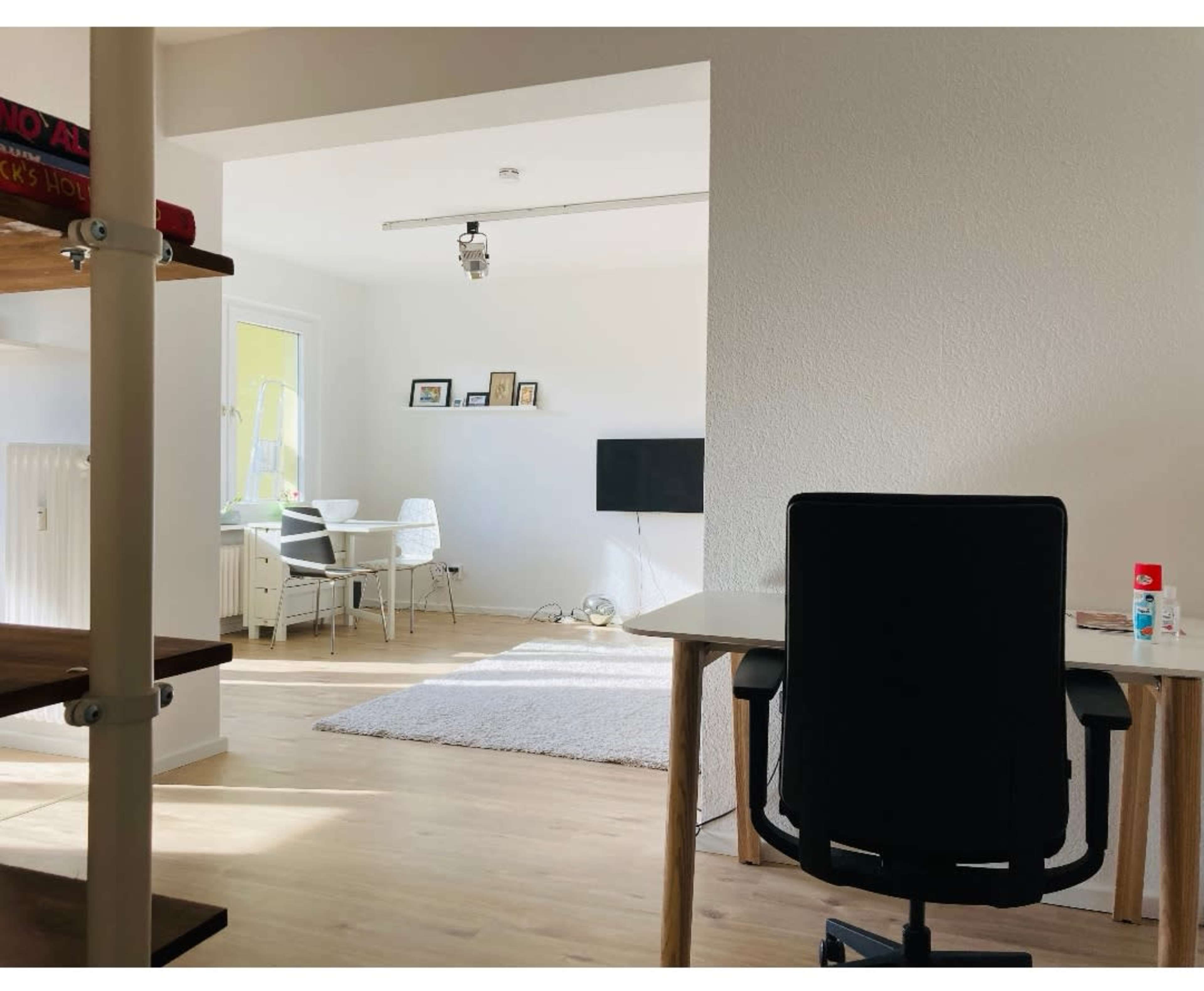 A spacious room features a desk and chair on one side, with a white carpet and seating area visible in the background.