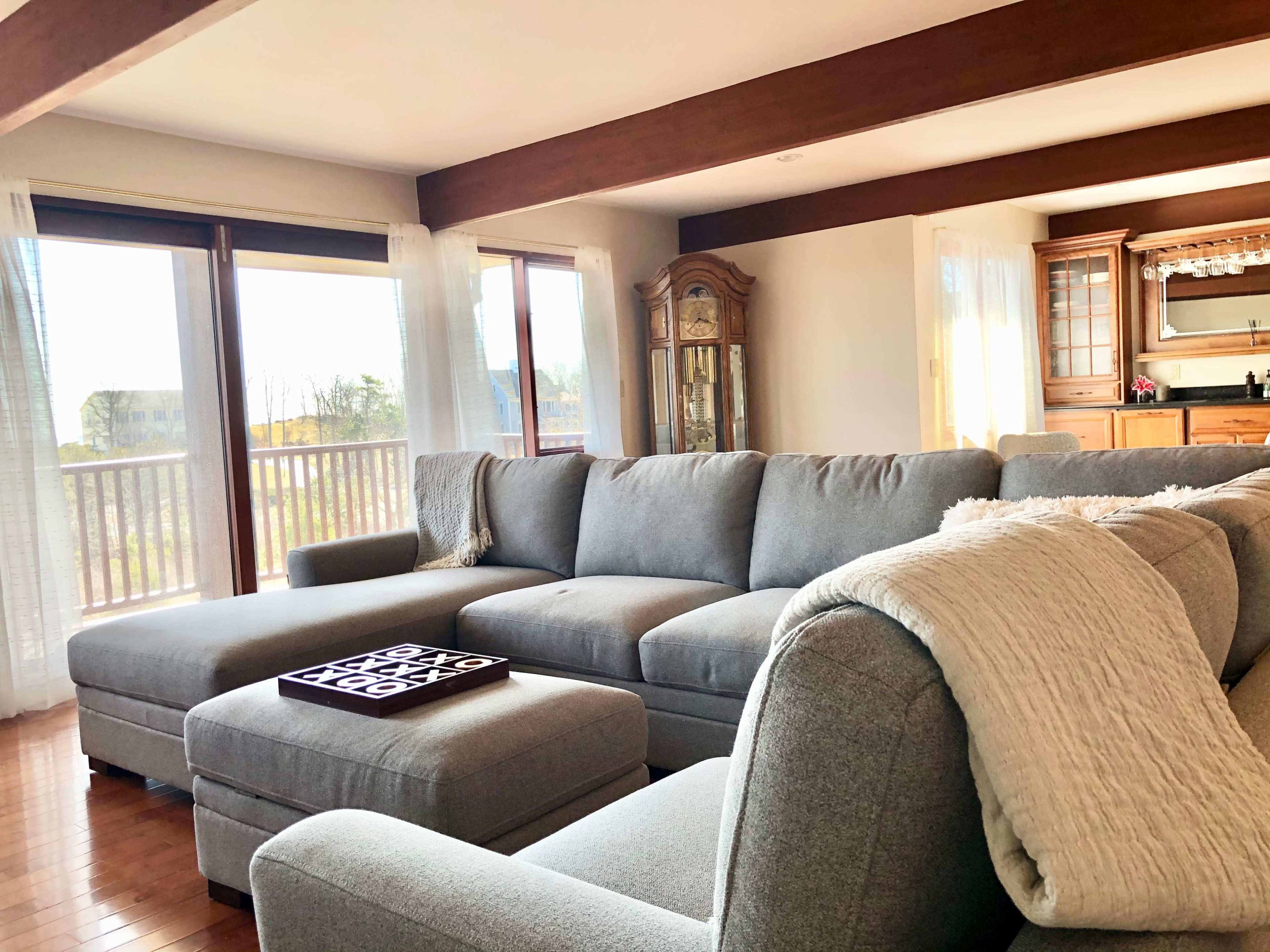 The living room features a large gray sectional sofa with a decorative throw blanket and a coffee table placed on a wooden floor, with windows providing natural light and a view of the outdoors.