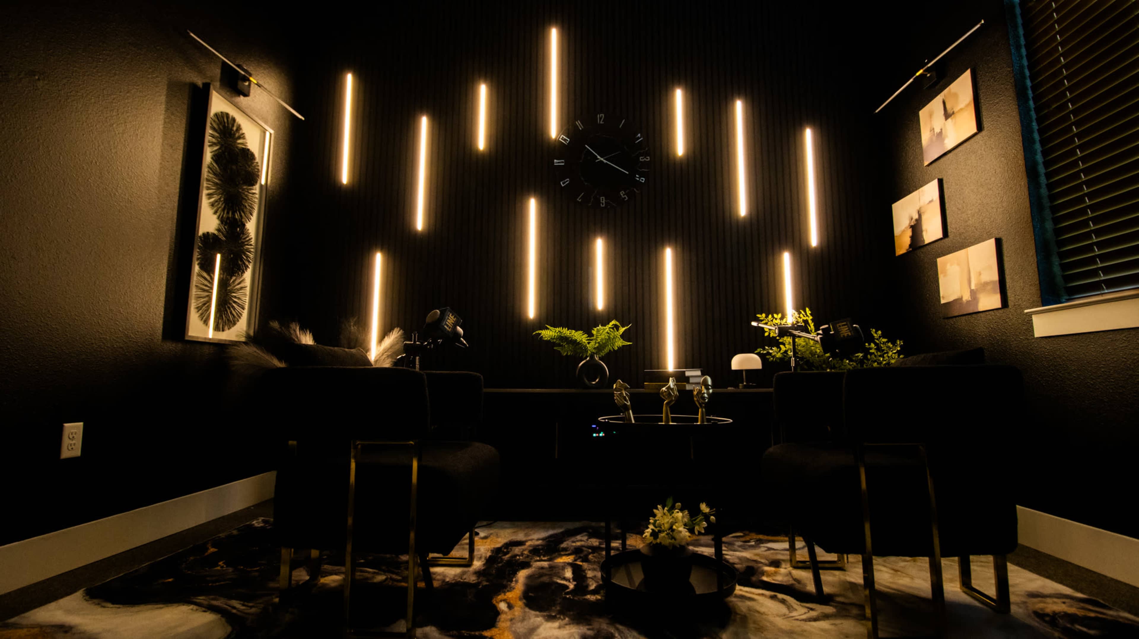 The image shows a modern, dimly-lit room with black walls, featuring illuminated vertical lines, a wall clock, and two black chairs positioned around a small table with plants and decor.