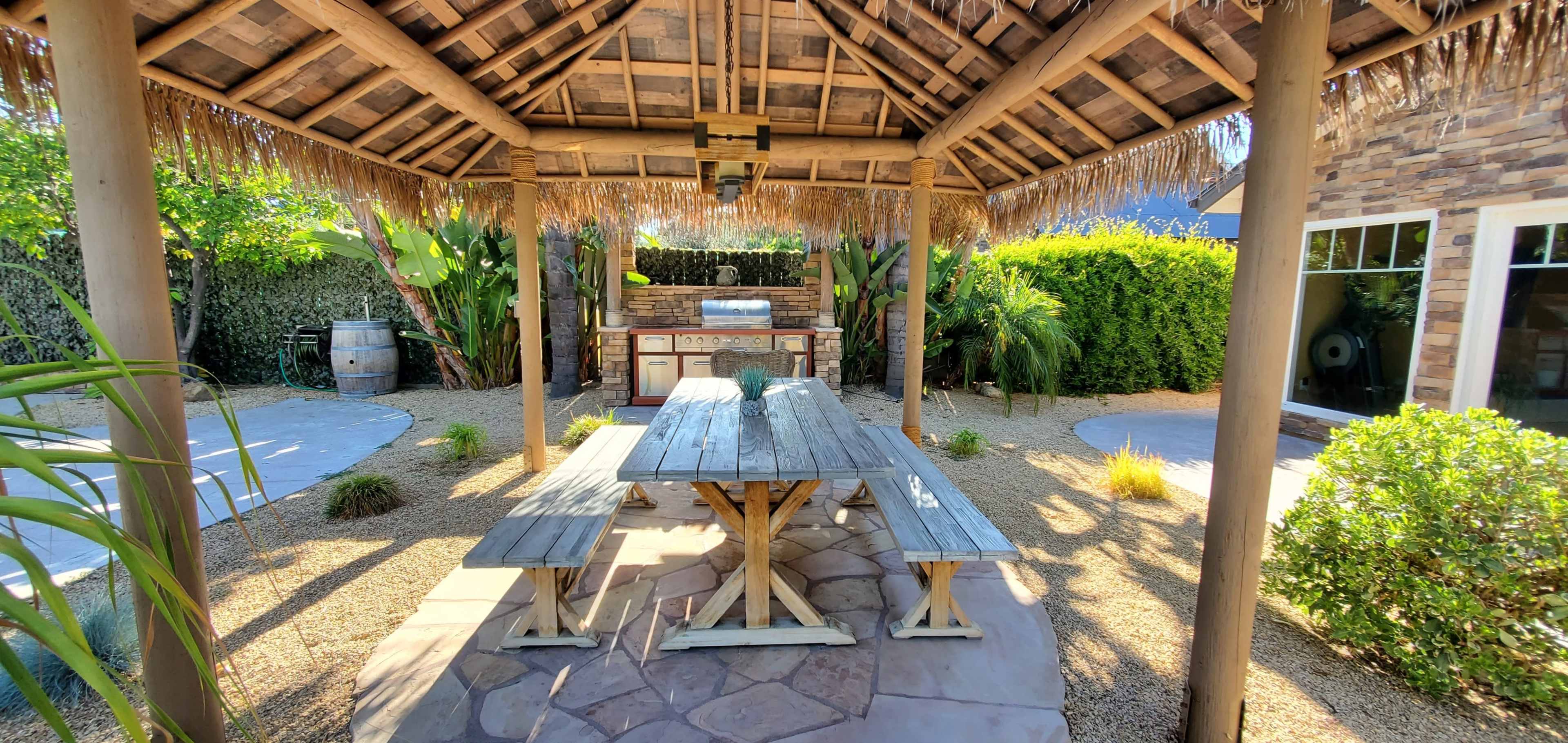 An outdoor patio with a thatched roof, a wooden picnic table, and an outdoor kitchen area surrounded by landscaping.