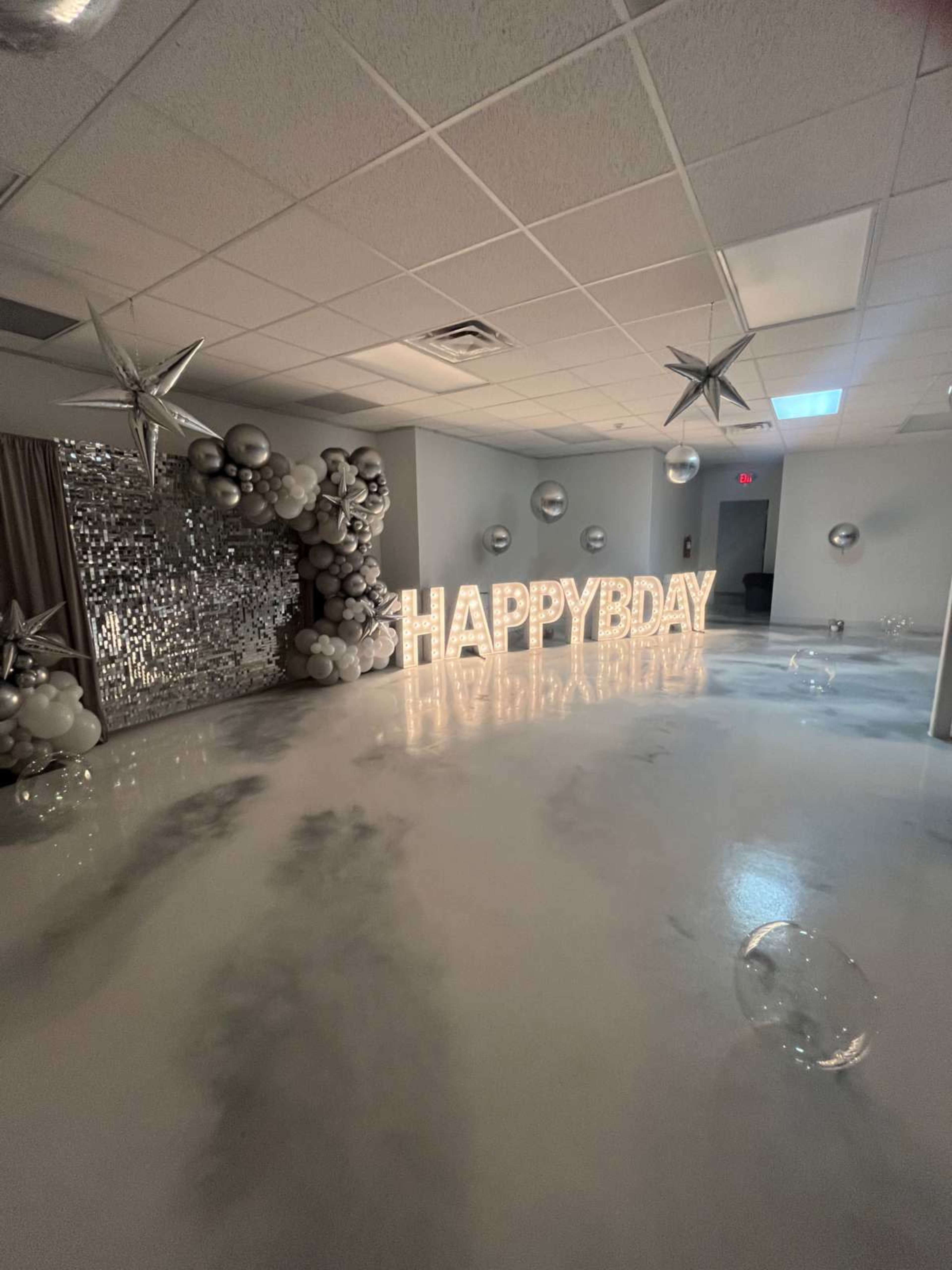 The room features a large "HAPPY BDAY" display illuminated by lights, surrounded by silver balloons and star decorations against a mirrored backdrop.