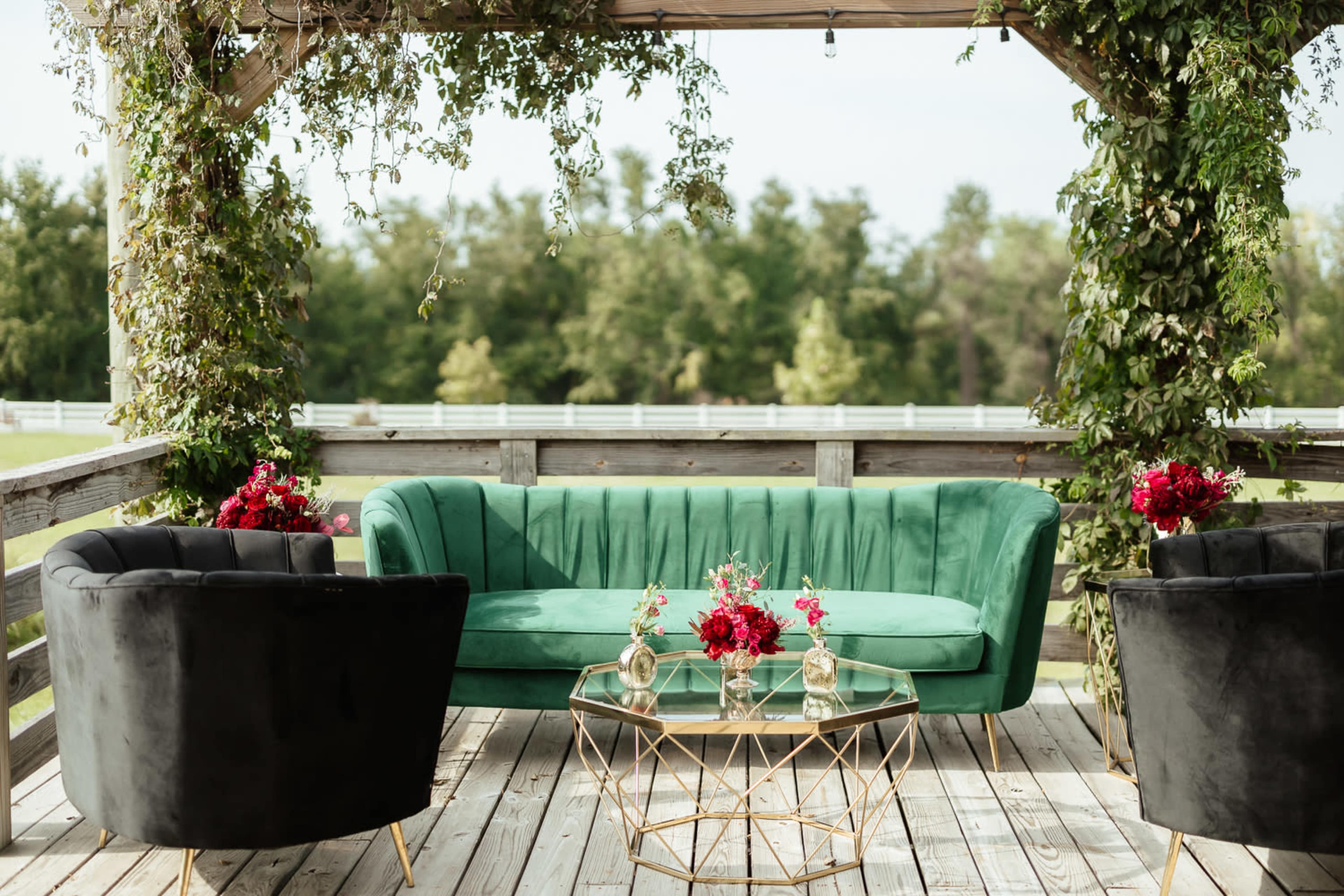 A green sofa with tufted upholstery is placed under a wooden gazebo adorned with vines, accompanied by two black chairs and a glass coffee table topped with floral arrangements.