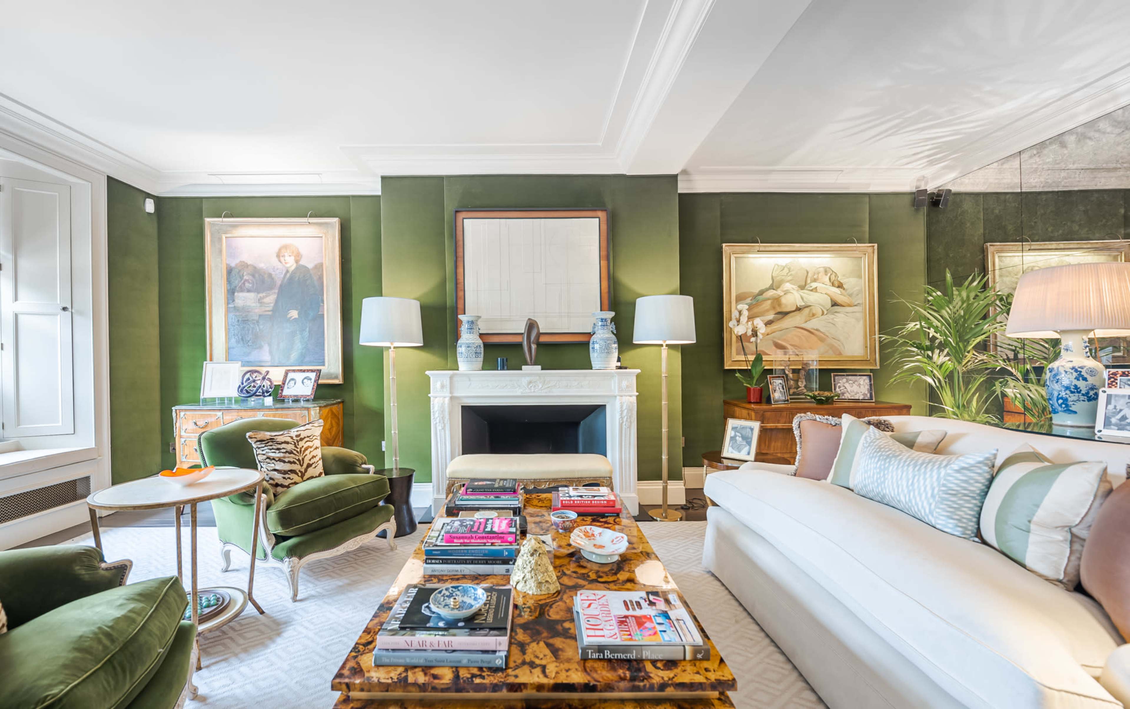 The living room features a green wall, a white fireplace with decorative art above it, and a mix of modern and antique furniture arranged around a coffee table stacked with books and decorative items.