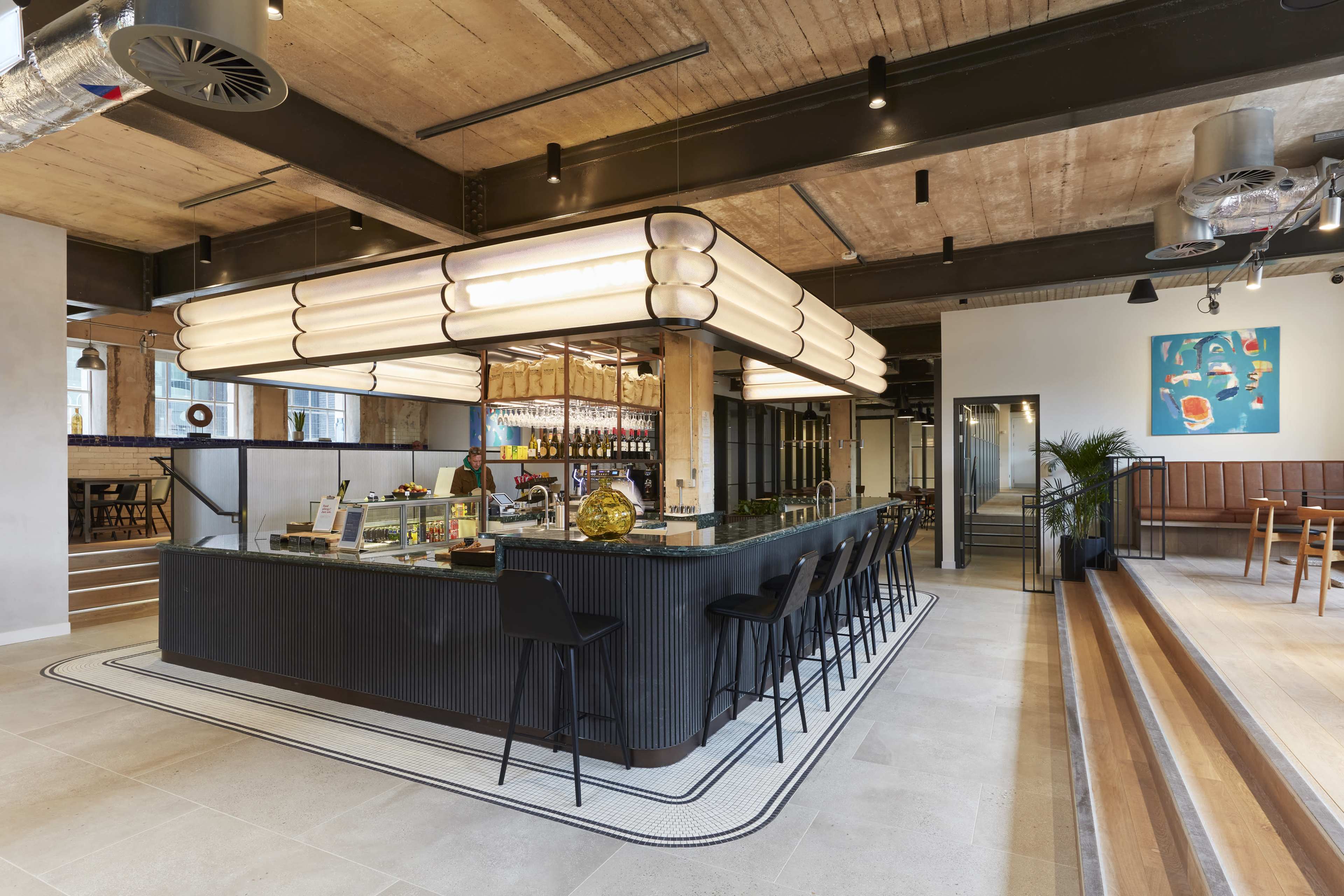 The image shows a modern café interior featuring a central black bar with seating, large pendant lights overhead, and minimalist décor.
