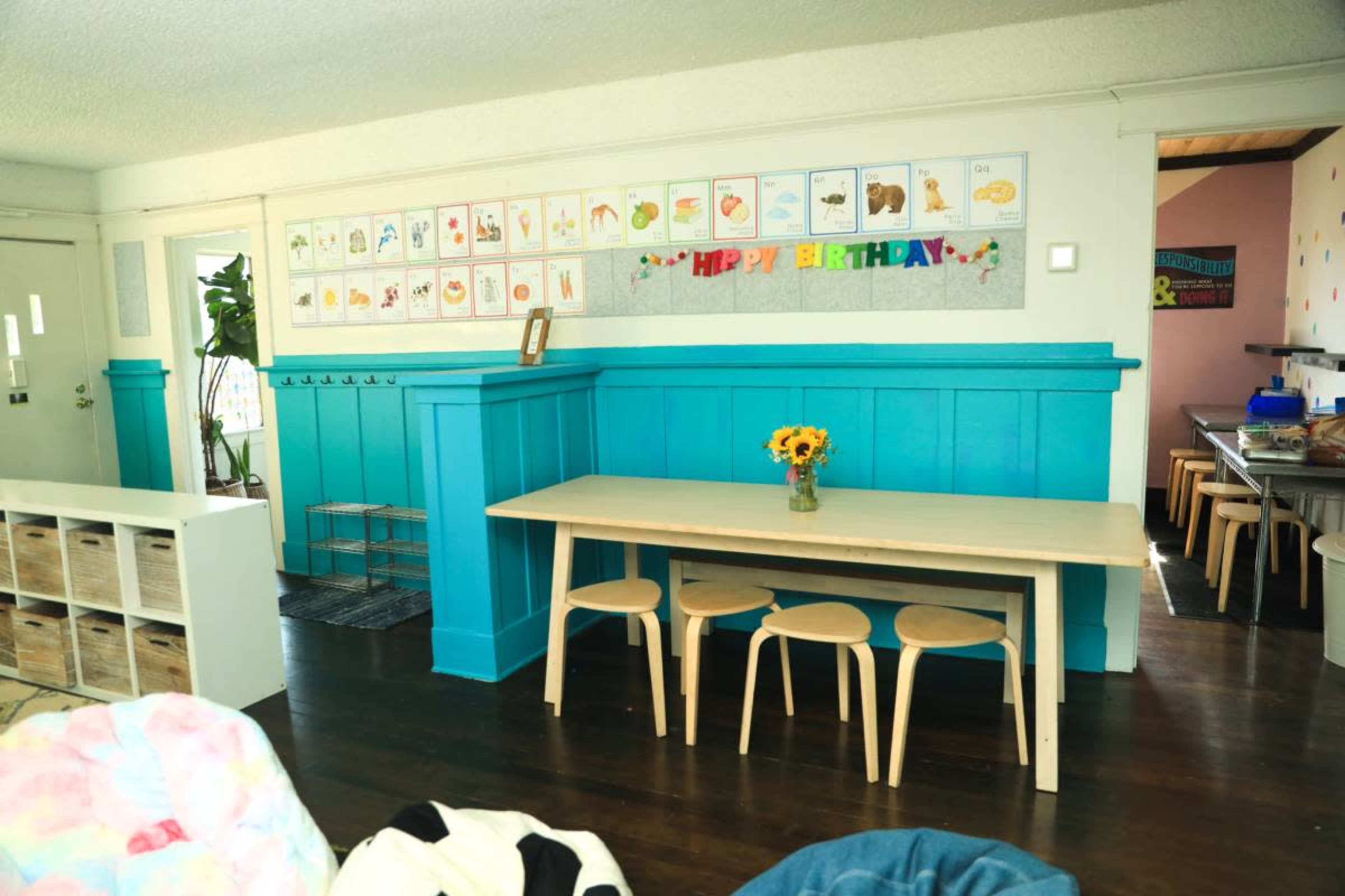 The image shows a brightly decorated room with turquoise walls, a simple wooden table surrounded by three chairs, and a flower vase as a centerpiece.