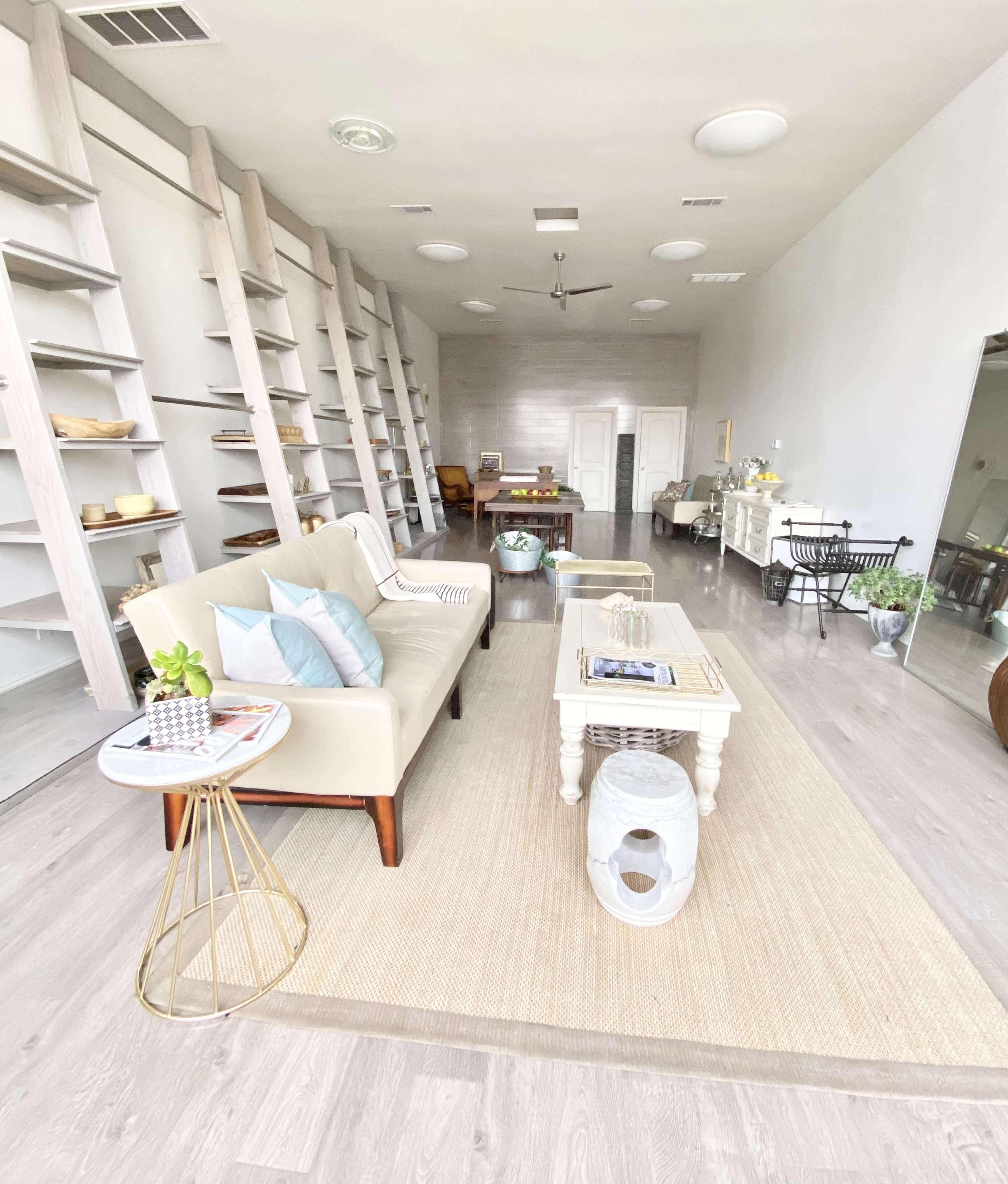 A spacious, modern living room with light-colored flooring, a beige sofa, a central coffee table, and shelves lining the walls.