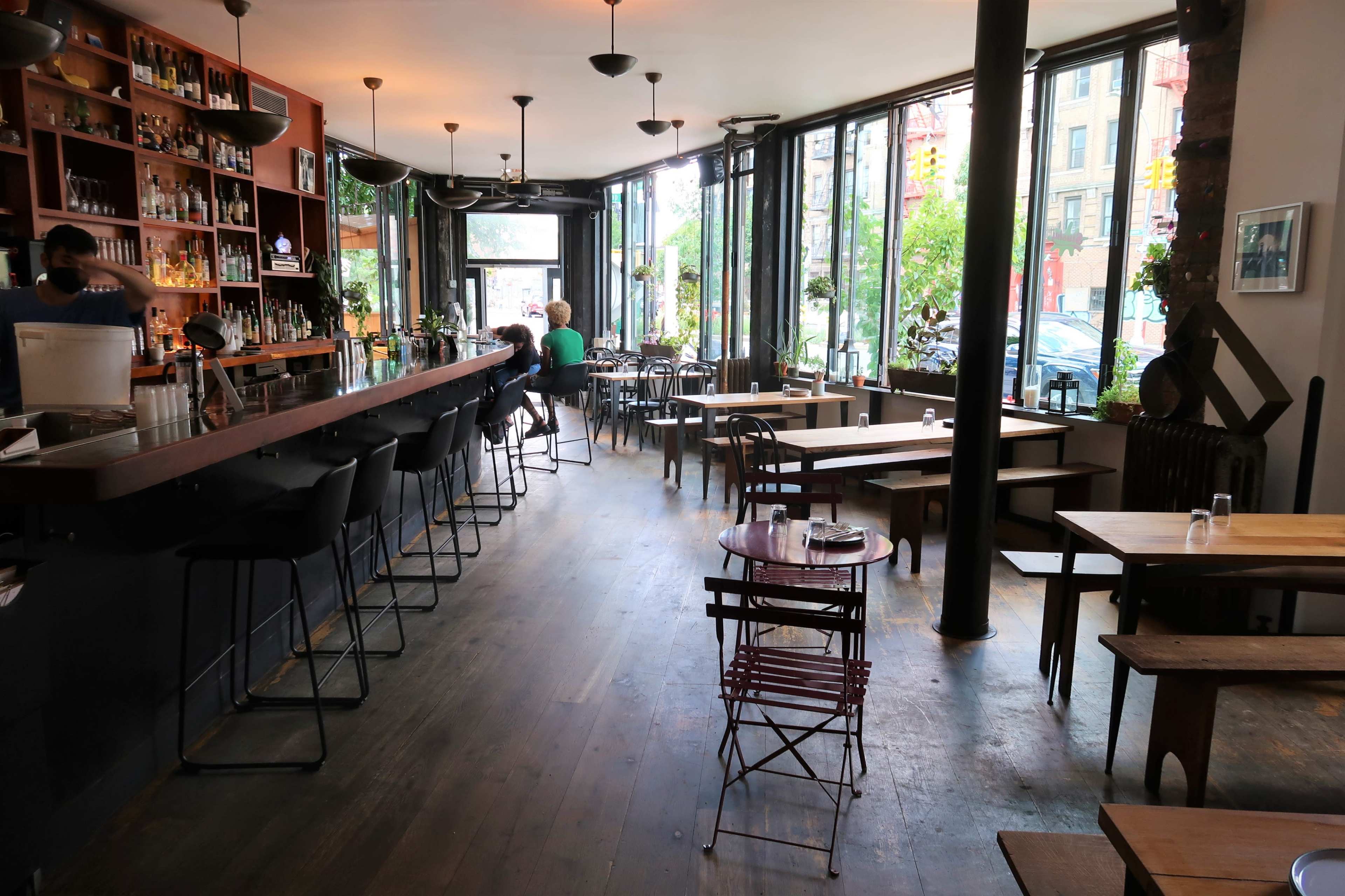 A restaurant interior with a long bar on one side, tall windows allowing natural light, and a variety of seating arrangements including tables and chairs.