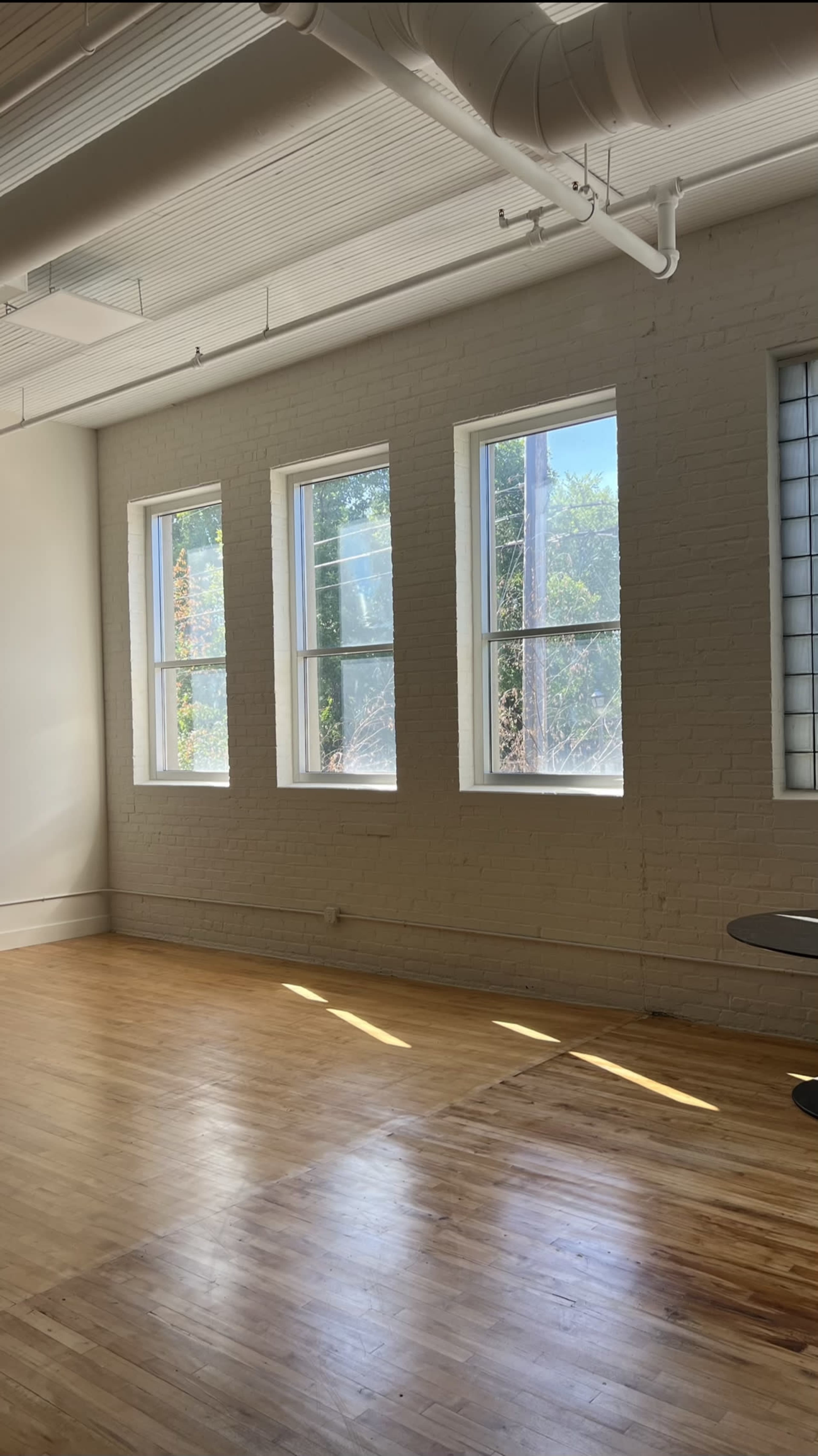 A spacious, empty room with three large windows allowing natural light to enter, illuminating the wooden floor.