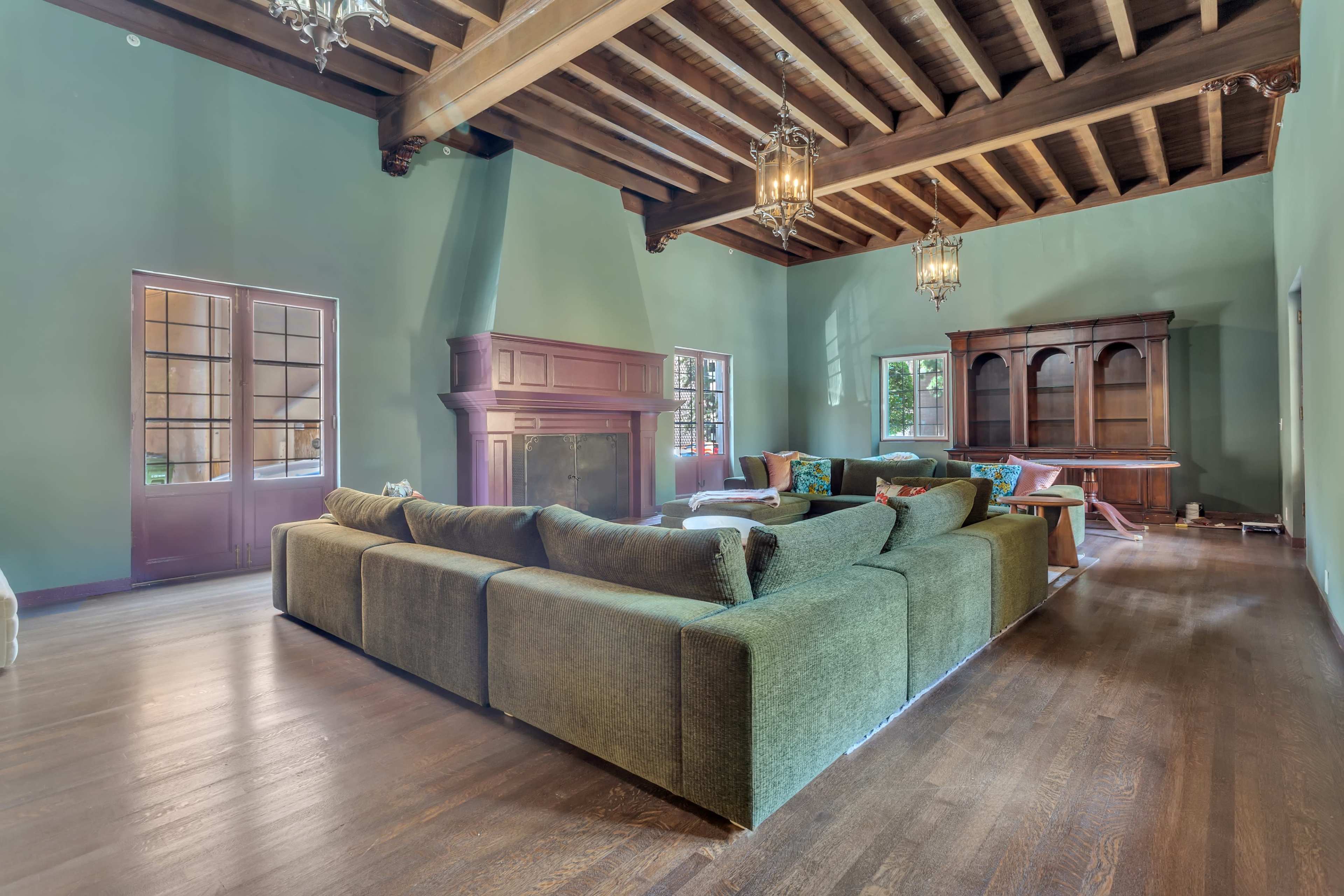 A spacious living room featuring a large green sectional sofa, wooden beams on the ceiling, and a fireplace with adjacent windows.