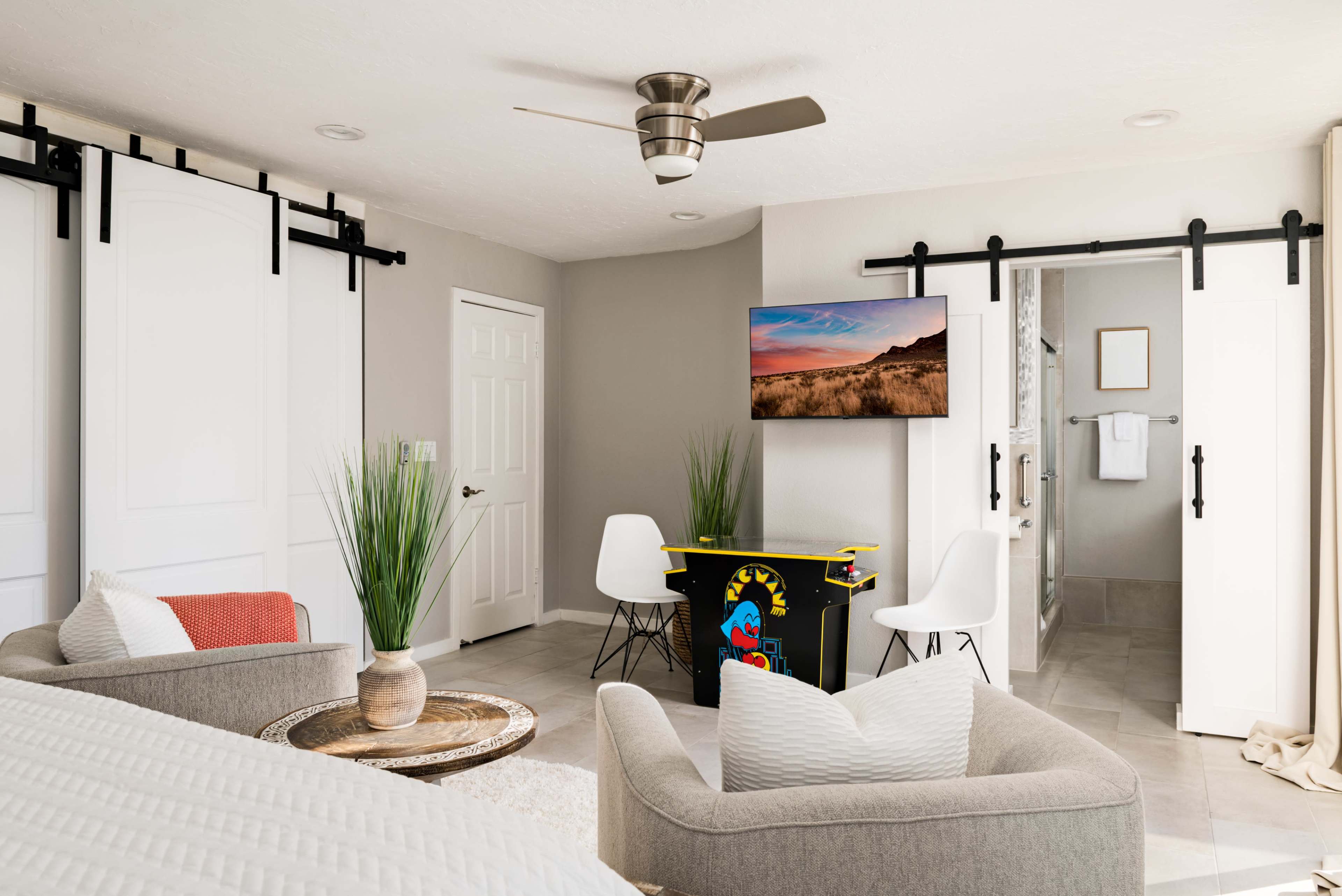 A cozy room features two chairs, a small gaming table, a mounted TV, and sliding barn doors, with neutral-colored walls and decorative plants.