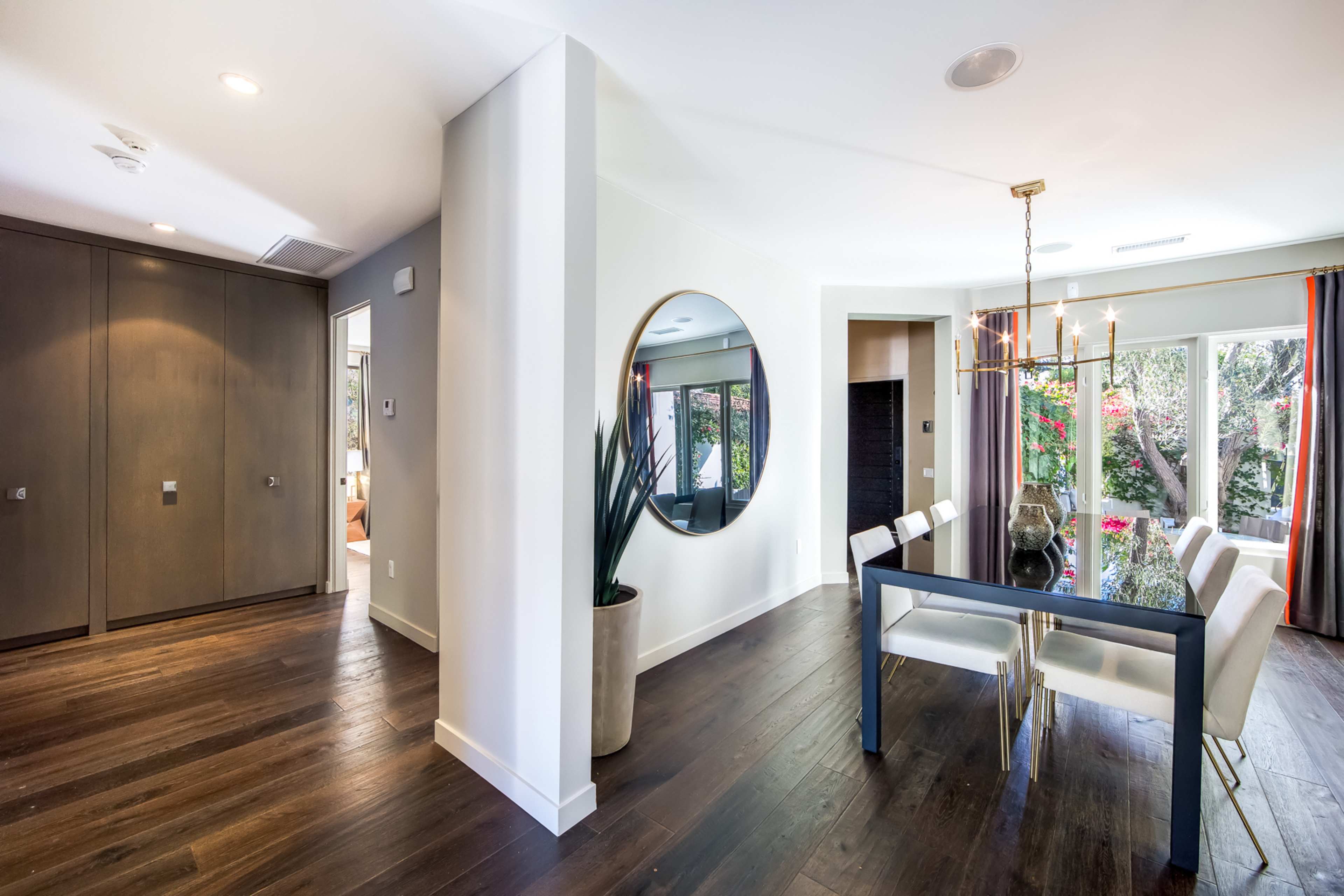 The image shows a modern dining area with a round mirror on the wall, a rectangular dining table, white chairs, and large windows overlooking greenery.