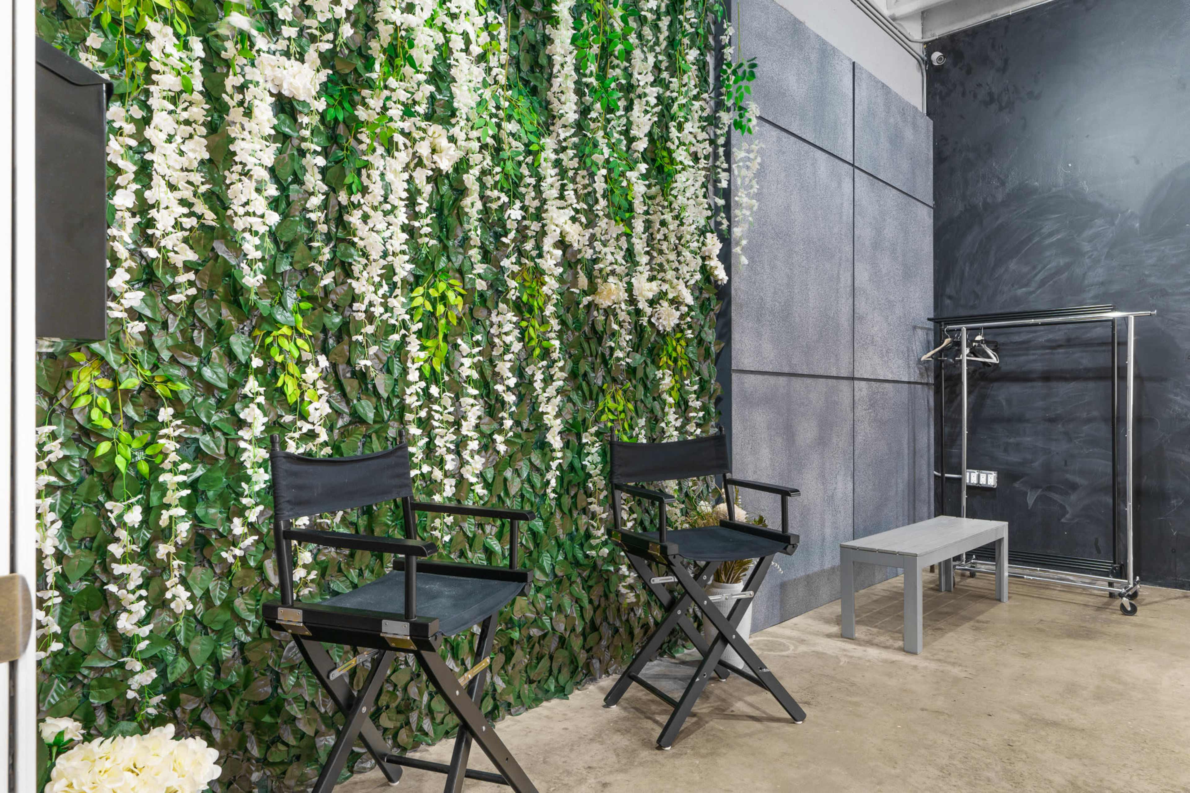 A room with a green wall covered in white flowers, two black director's chairs, and a gray bench in front of a clothing rack.
