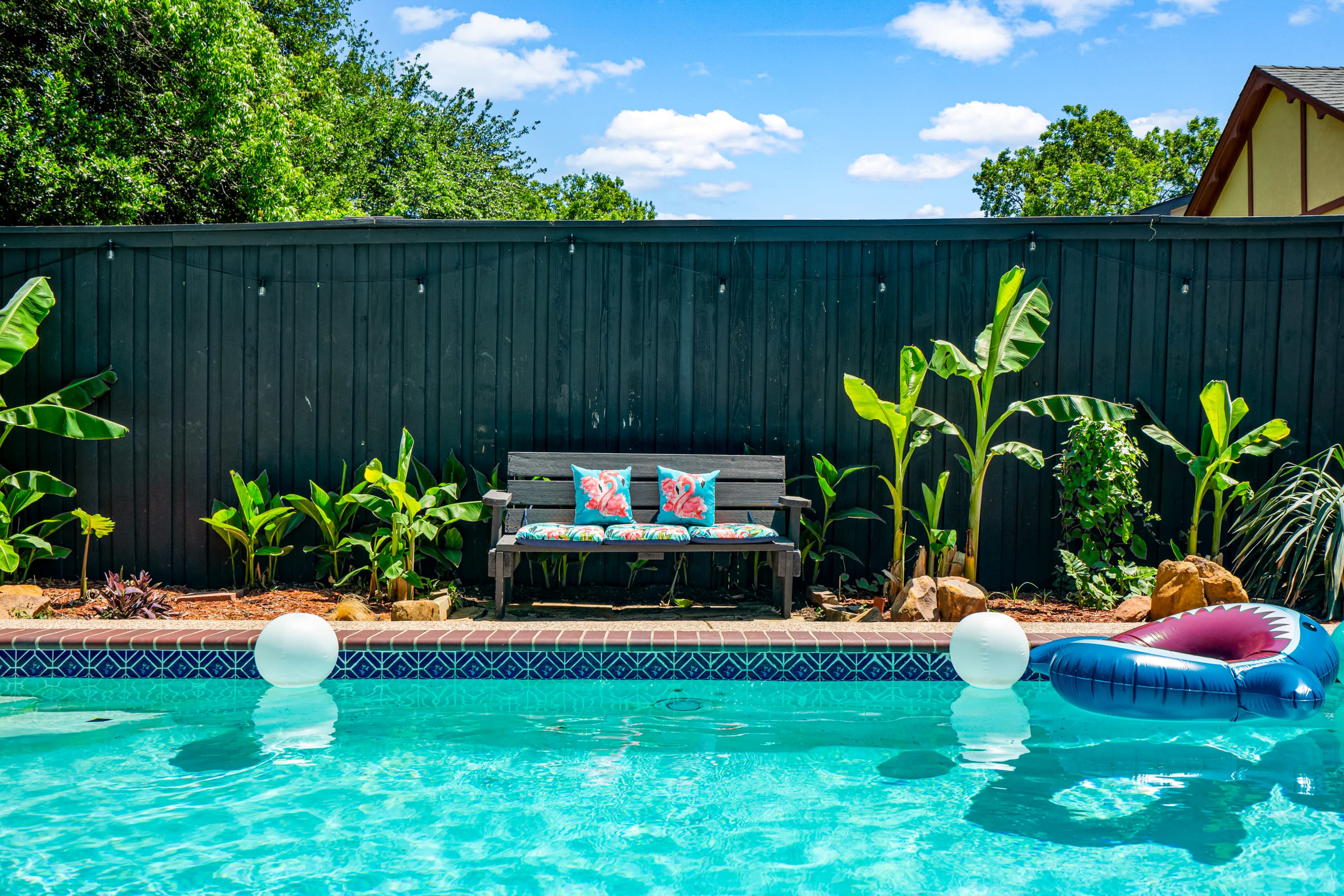 A wooden bench with colorful cushions is positioned beside a swimming pool, surrounded by greenery and inflatable floaties.