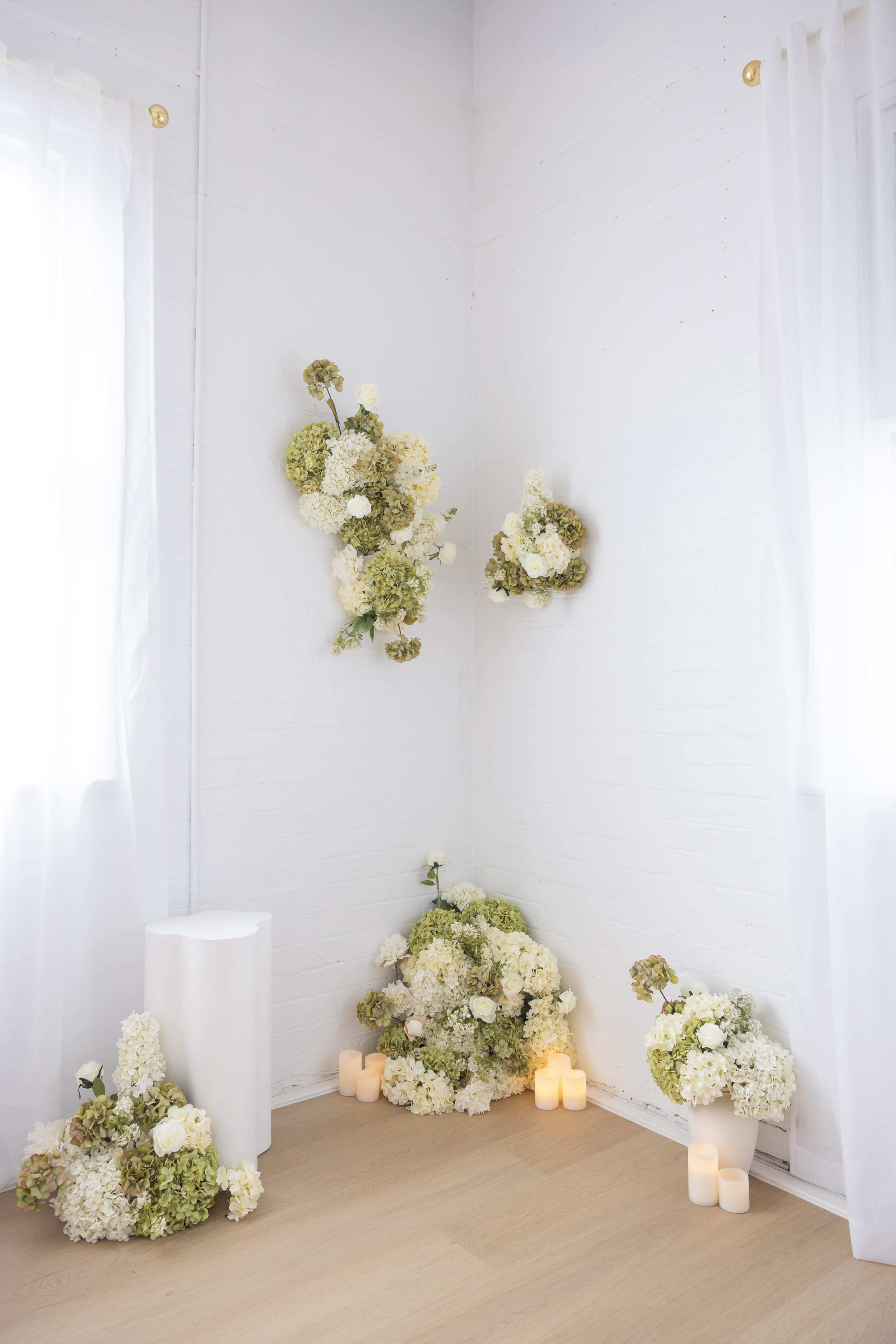 A corner of a room features white and green floral arrangements on the walls and floor, accompanied by lit candles.