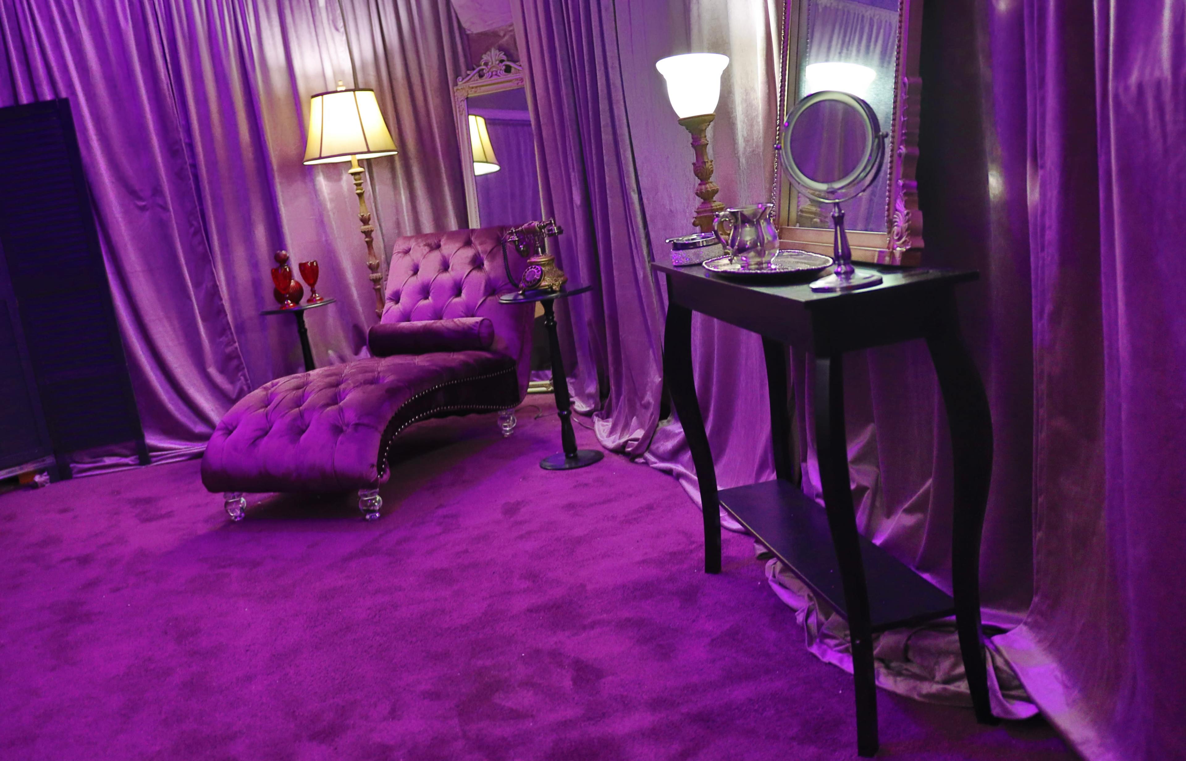 A purple-themed room featuring a chaise lounge, a table with a mirror and toiletries, and decorative lamps.