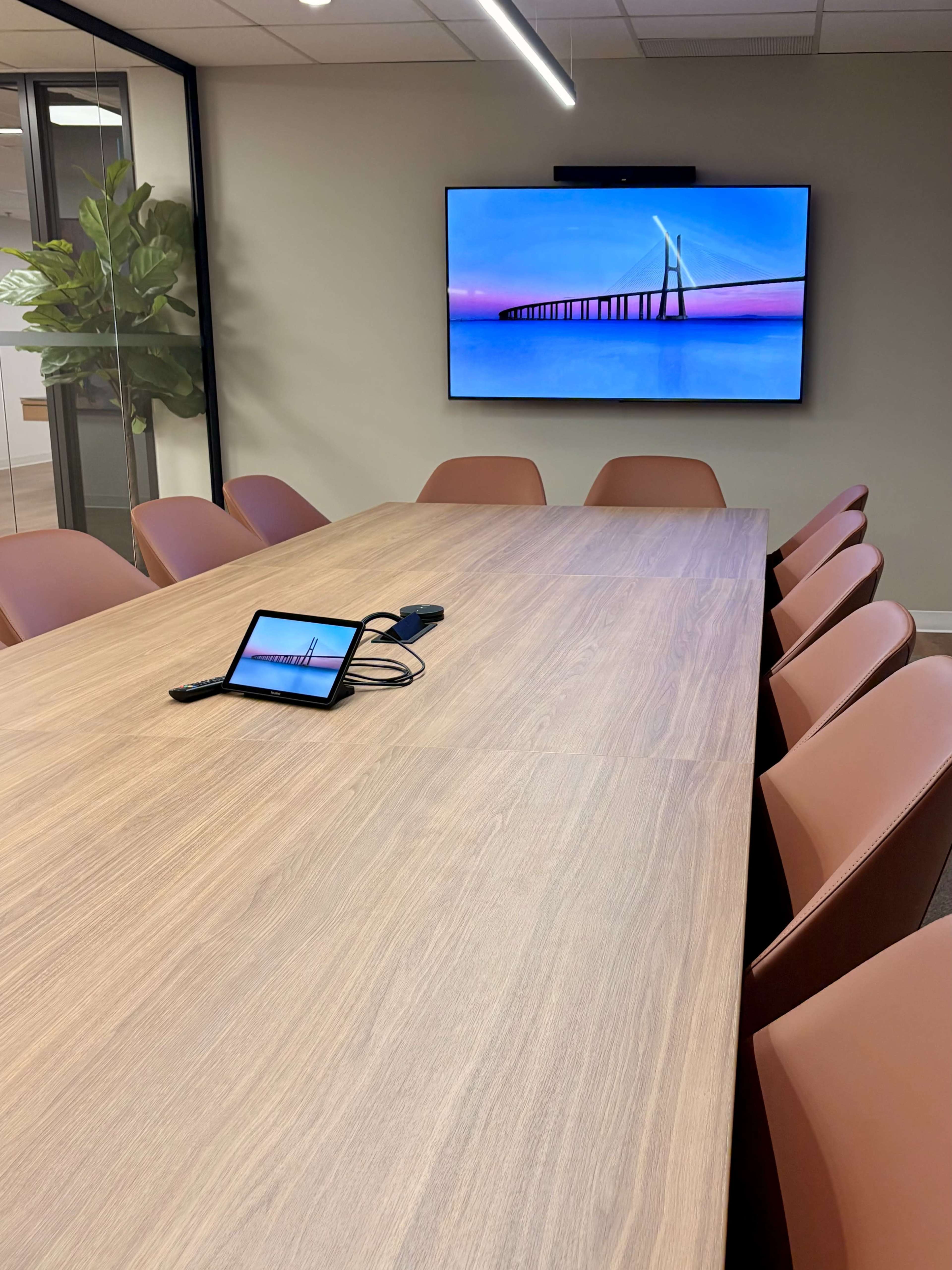A modern conference room features a long wooden table surrounded by brown chairs, with a large screen displaying a scenic image on the wall.